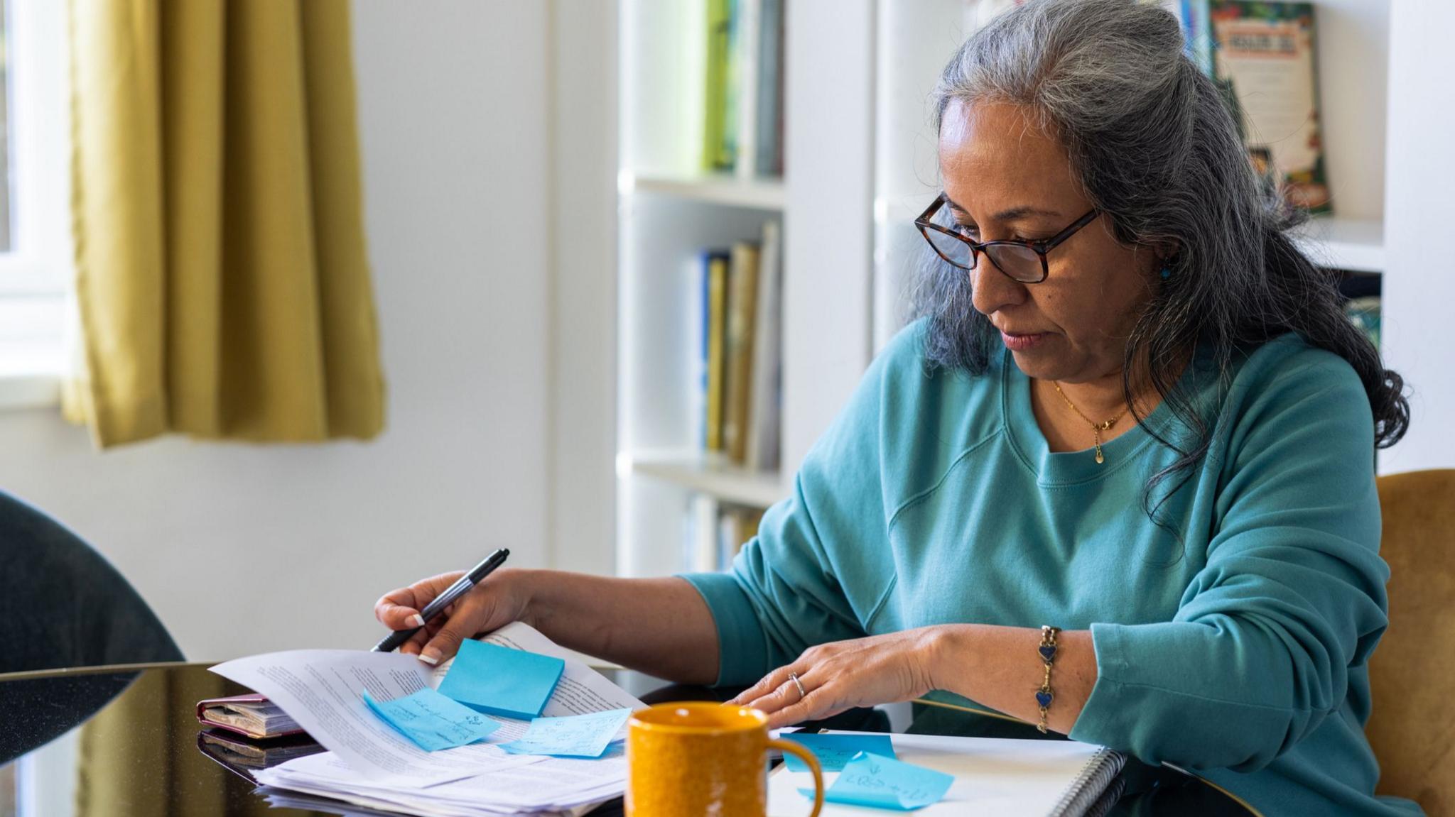 A woman sits at a table, holding a pen in one hand as she looks down to a stack of papers and post-it notes