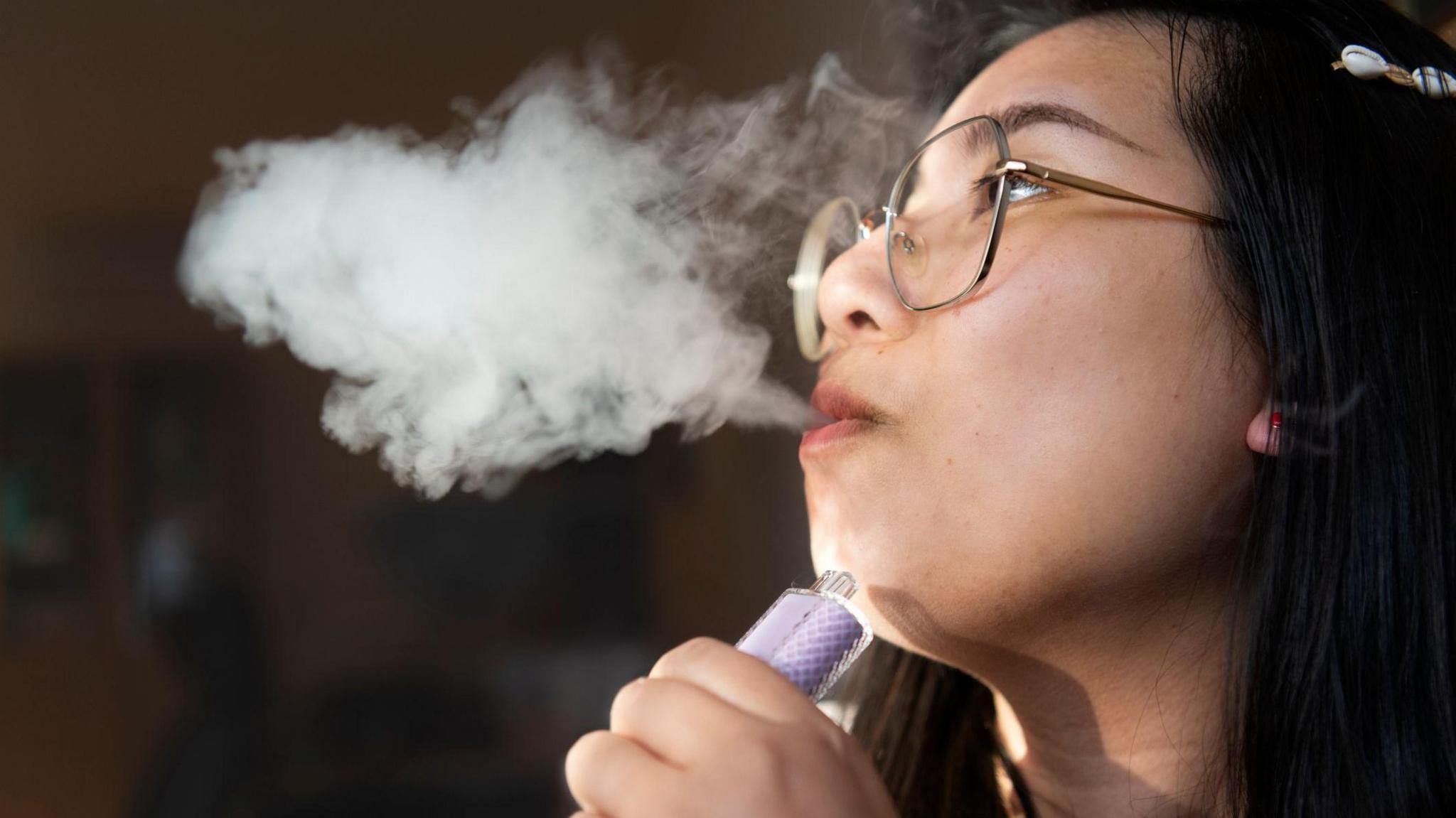 Young woman with large metal framed glasses, vaping in a darkly lit room. There is a large cloud of smoke from her mouth. 