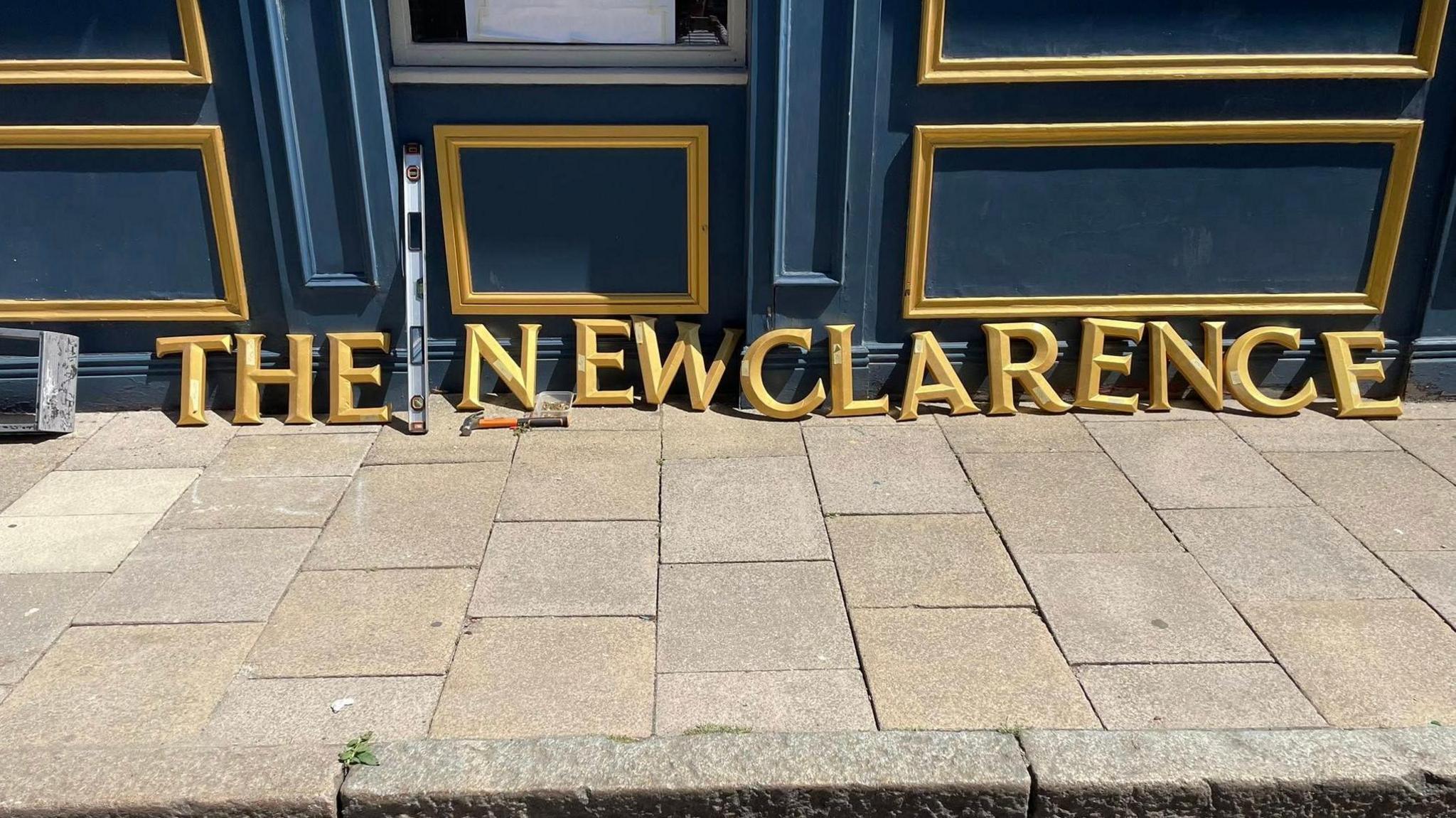 Blue pub facia with gold lettering resting on the pavement reading 'The New Clarence'.