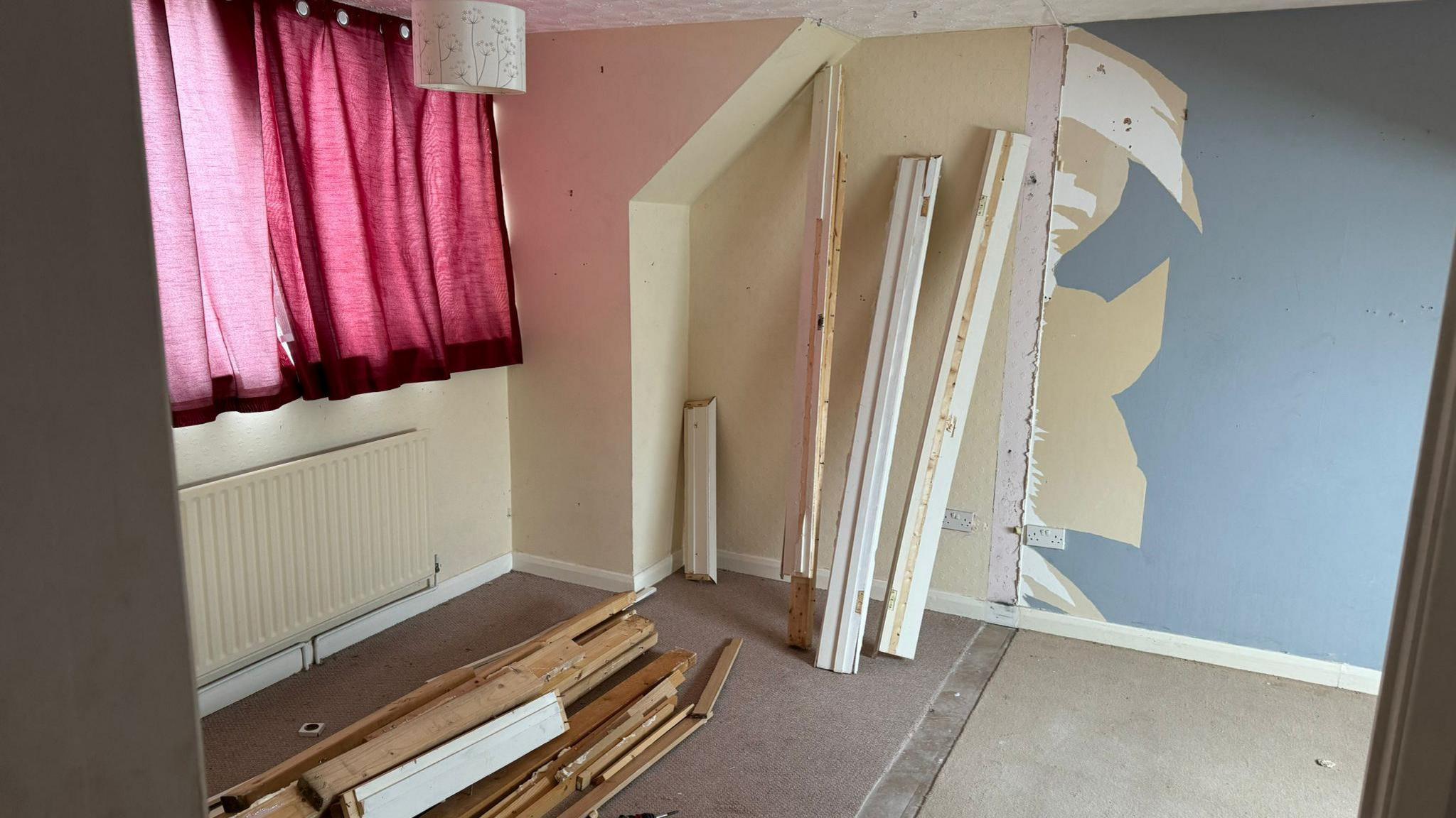 A room in a house where the wall appears to have been removed. Panels of wood rest of the floor and the wallpaper has been partially ripped off the wall. The pink curtains in the room are drawn. 
