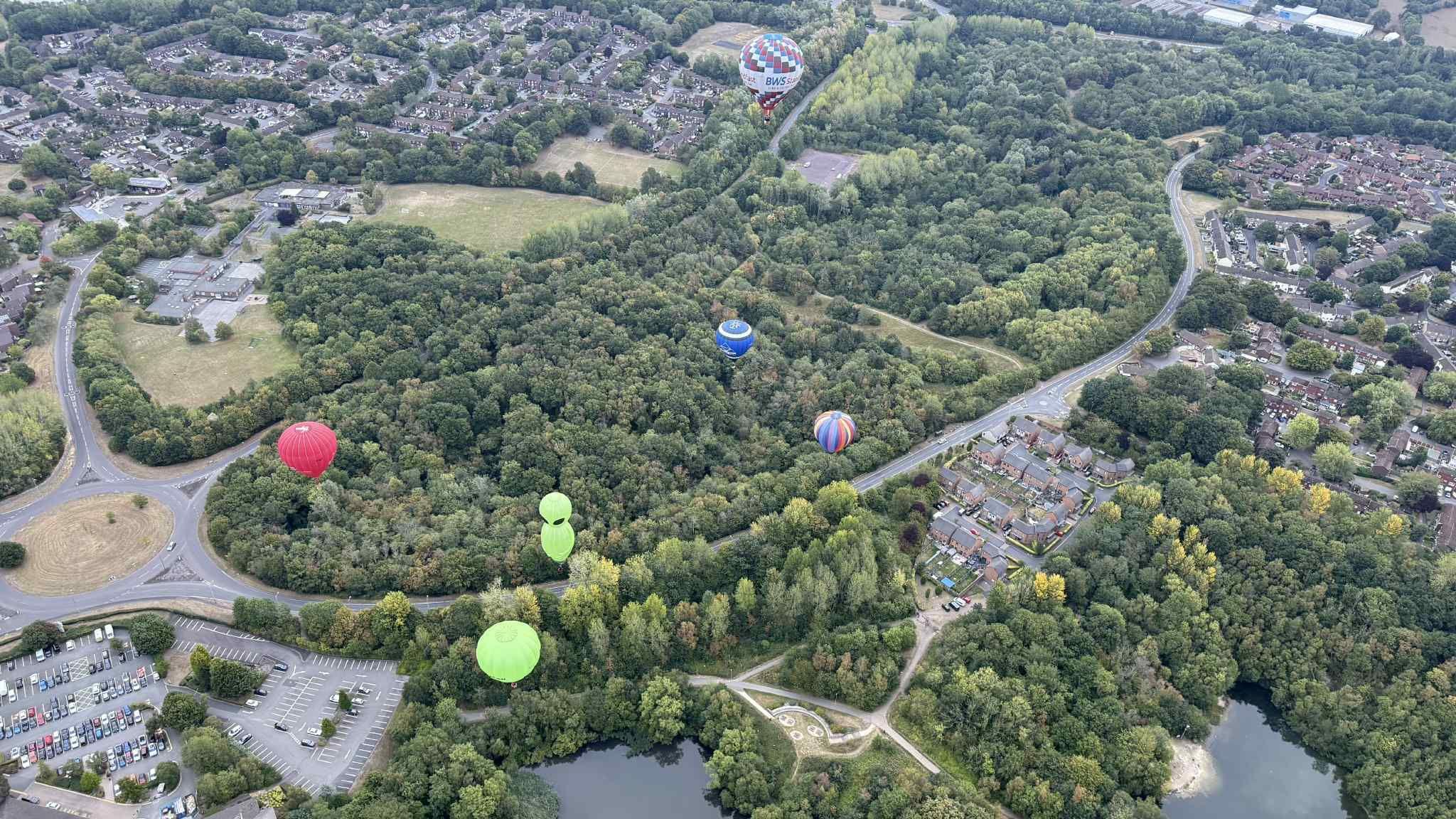 A birds eye view of multi-coloured hot air balloons in the sky while trees, a car park and buildings are below.