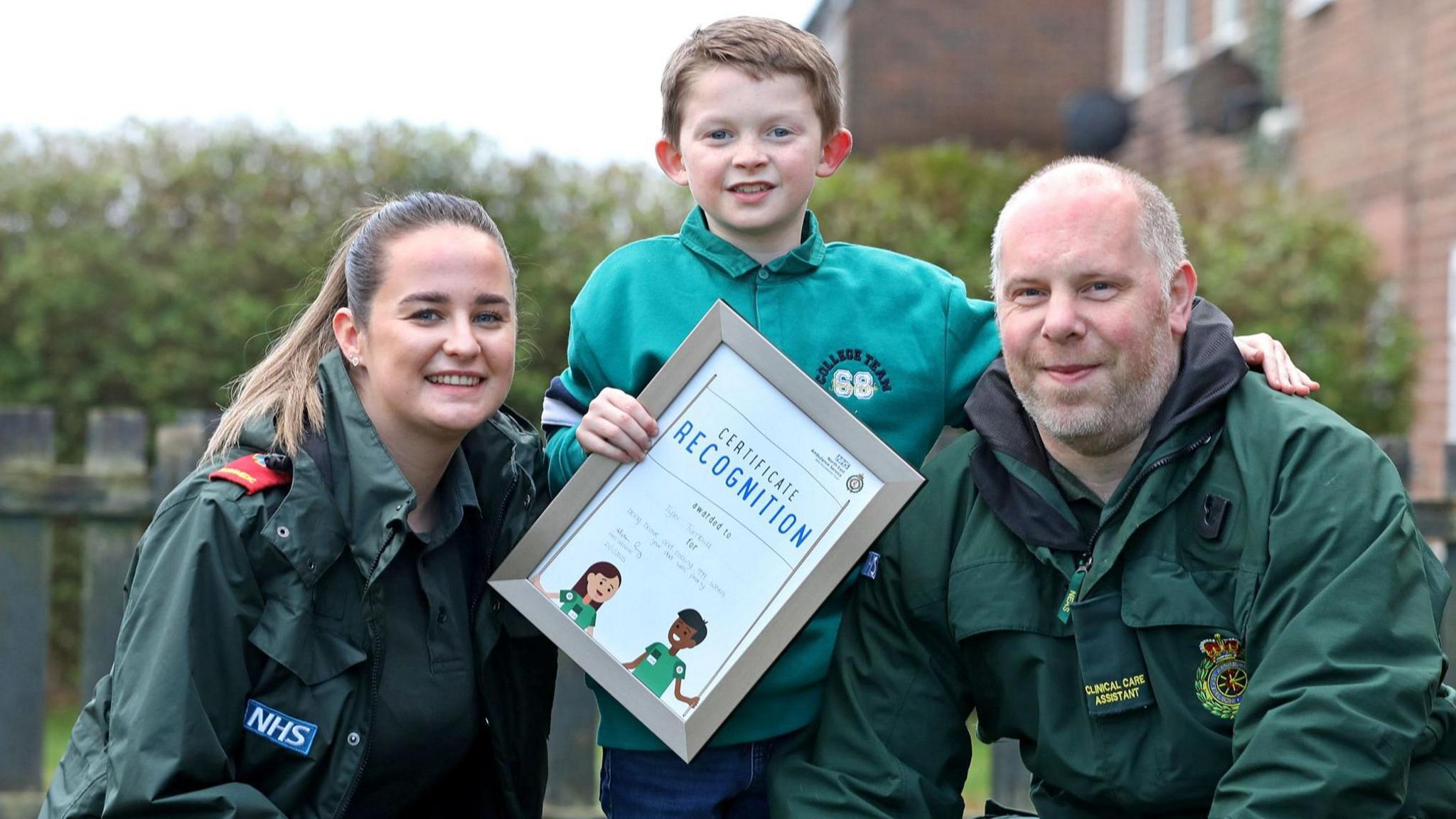 Tyler, who is eight years old, stood beside two emergency care assistants, one man and one woman. They are pictured smiling in green NHS work uniform. Tyler, in a blue sweatshirt, is pictured holding his certificate of recognition for the call he made to 999 during his father's diabetic attack.