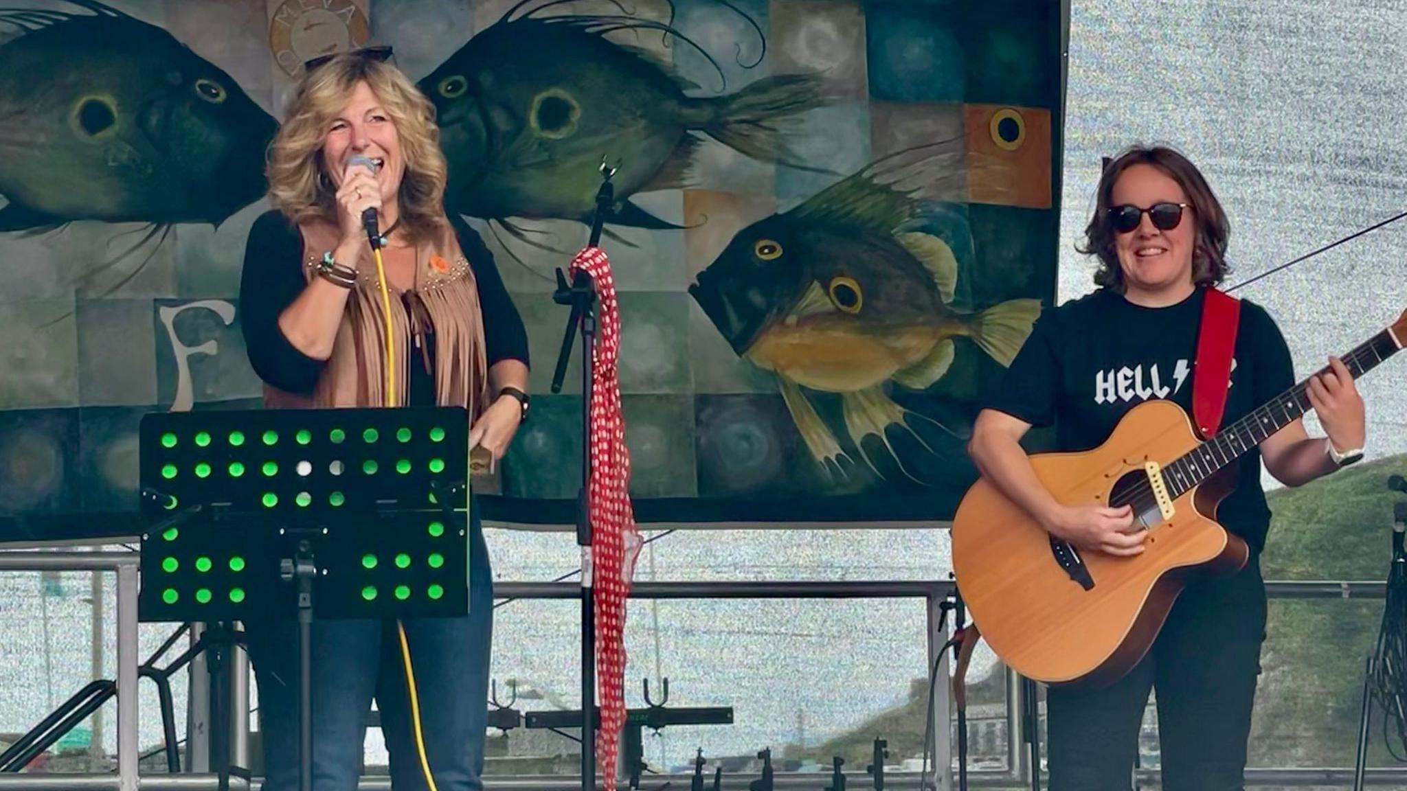 Two women perform music on a stage. The woman on the left is singing into a microphone. The woman on the right is playing an acoustic guitar which is strapped over her shoulder.