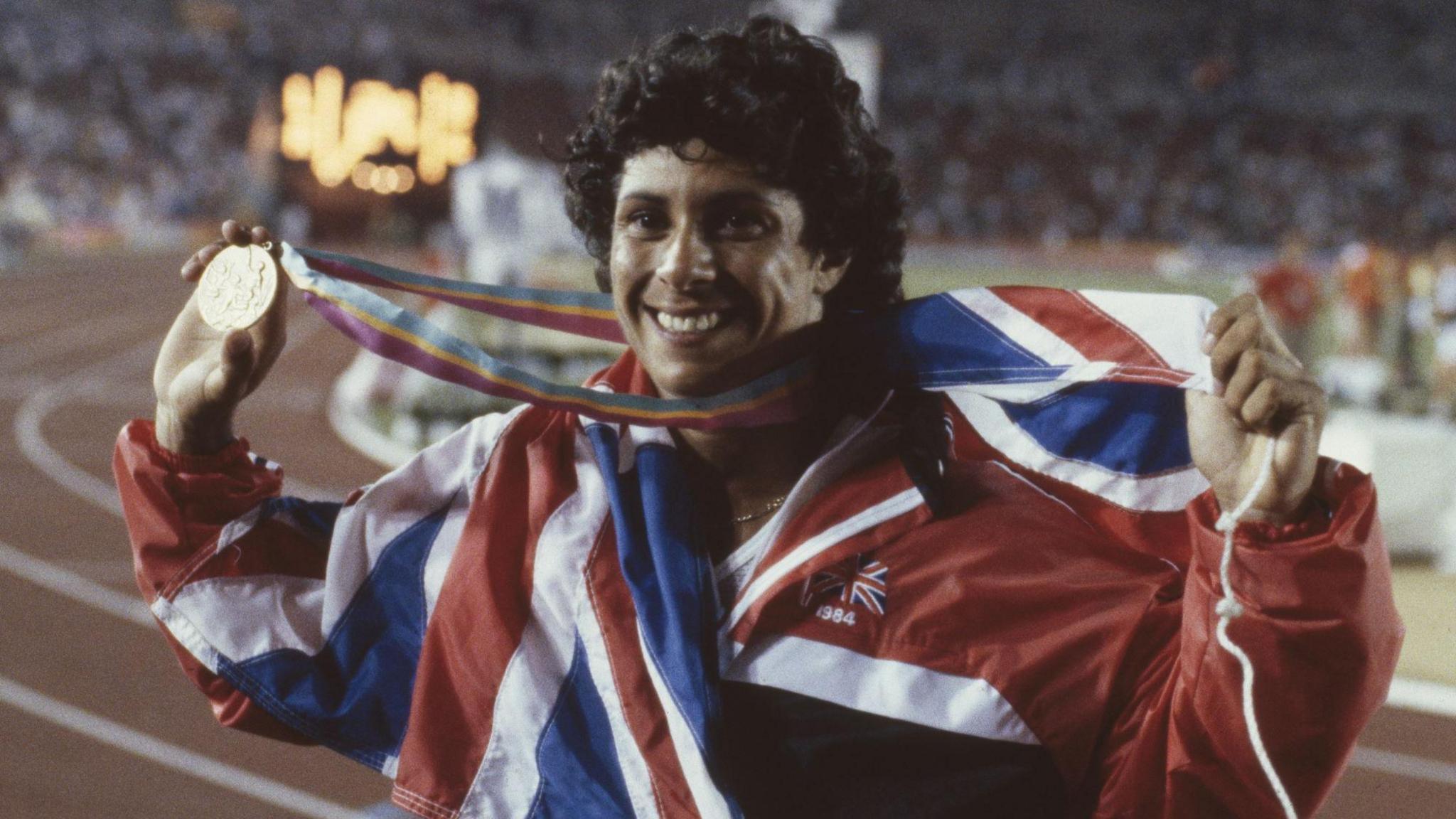Fatima Whitbread stands on an athletics track holding her bronze medal up for the stadium audience to view. She has the Union Jack flag draped around her shoulders and is smiling.
