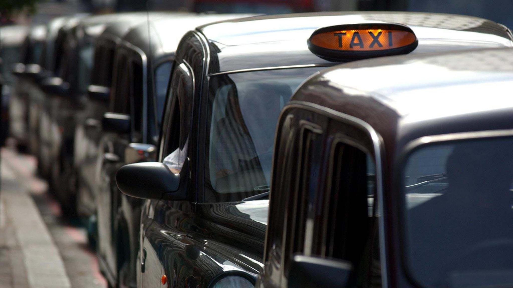 A row of black taxis with one car having an illuminated orange taxi sign