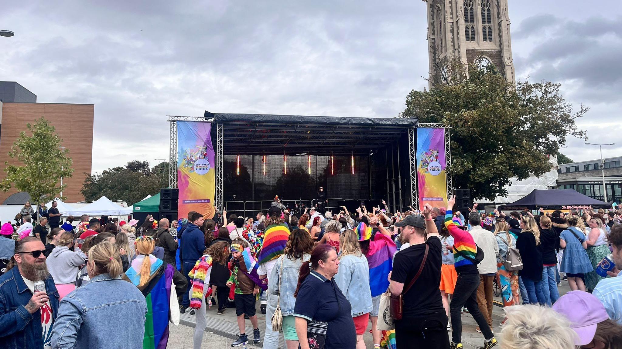 A crowd of people stand in front of the stage at Scunthorpe Pride in the town centre, many are wearing rainbow pride flags and clothing.