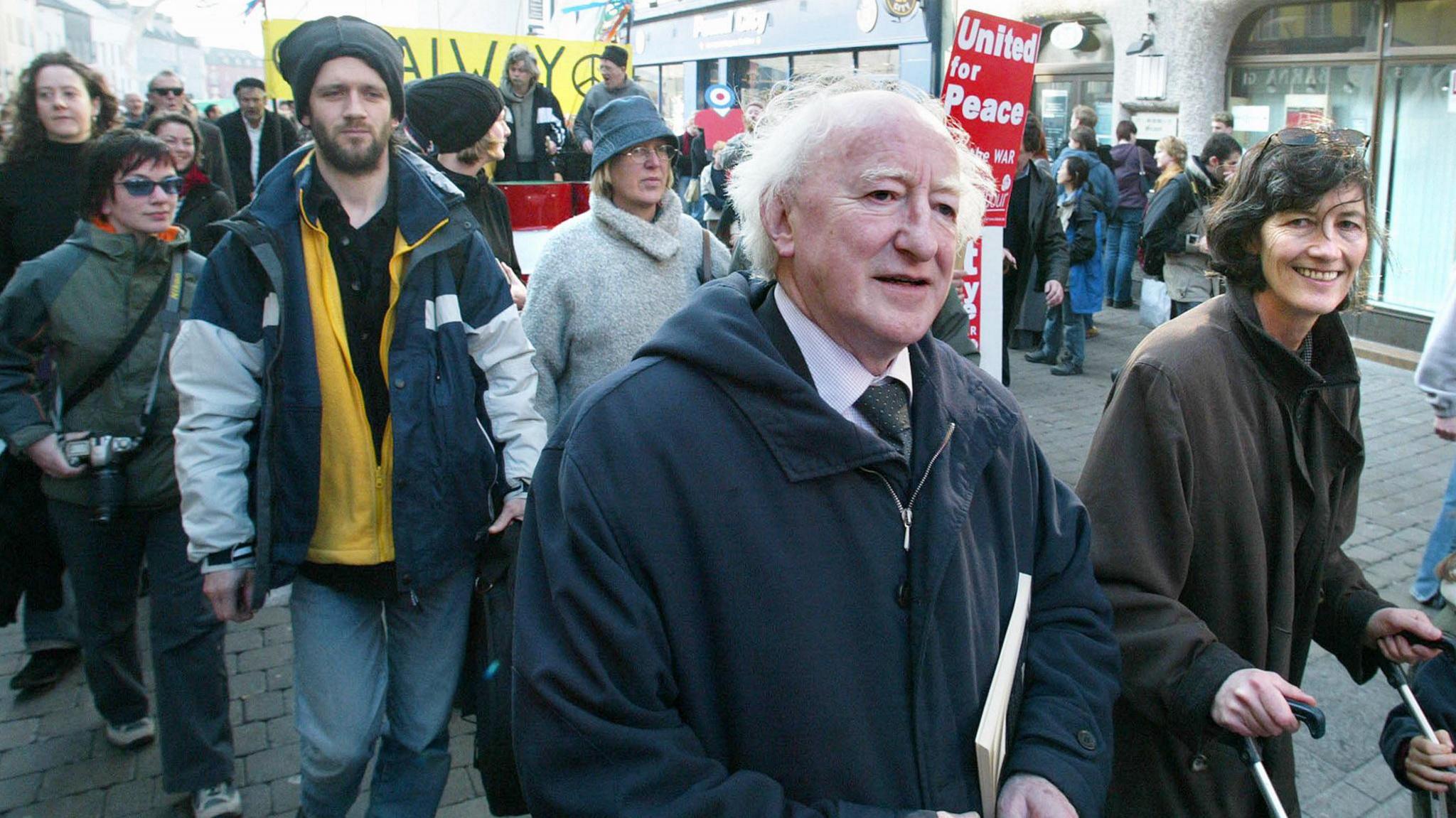 Catherine Connolly and Michael D Higgins marching on a street in Galway in 2003.  Connolly is smiling and pushing a child's buggy. She is wearing a long brown coat and has sunglasses on her head. Higgins has white hair and is wearing a navy coat over a suit.  A crowd of fellow protesters are following behind them.