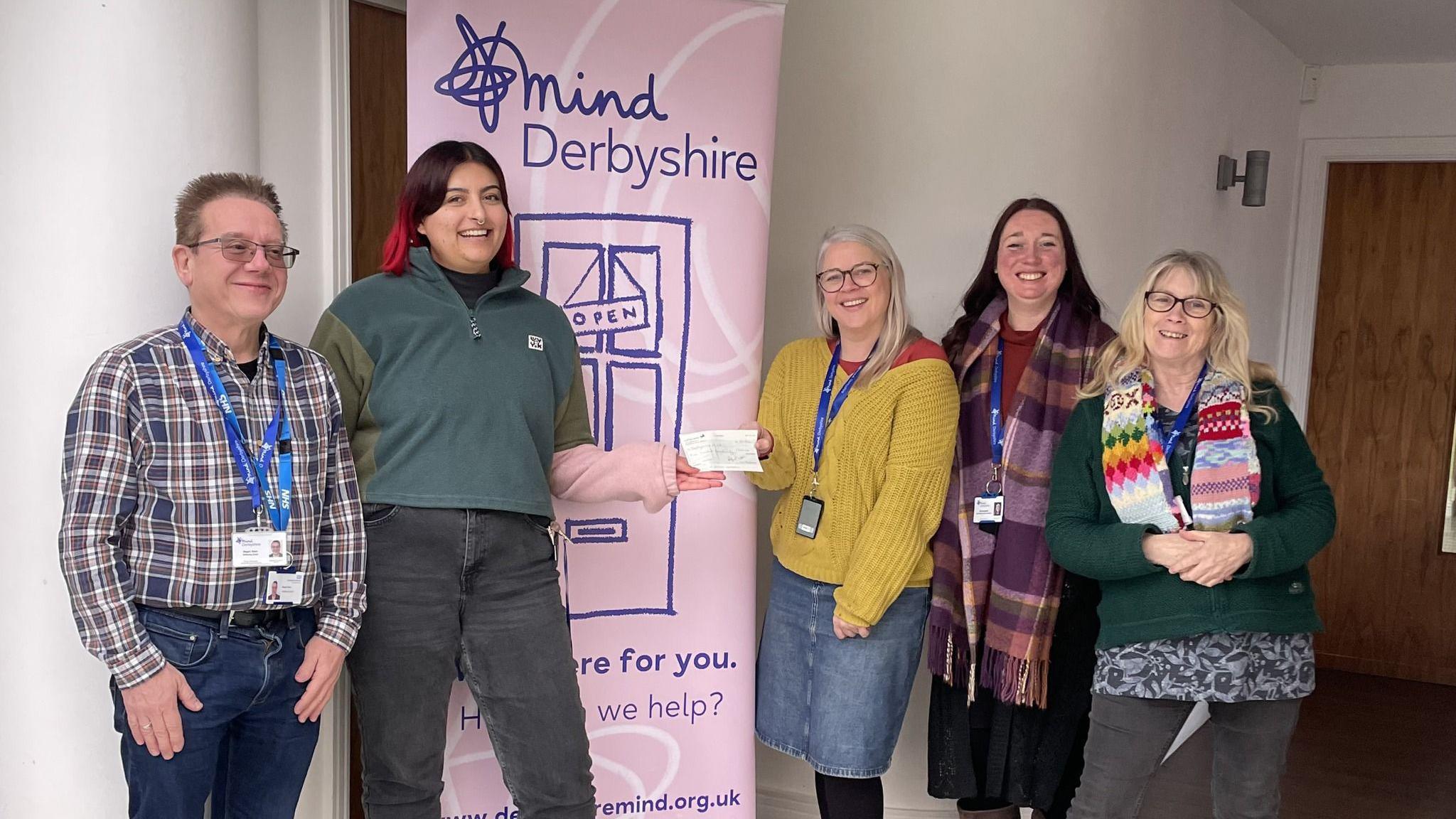 Five people stood by a pink sign saying "Mind Derbyshire" with a cheque being handed over from one woman to another. 