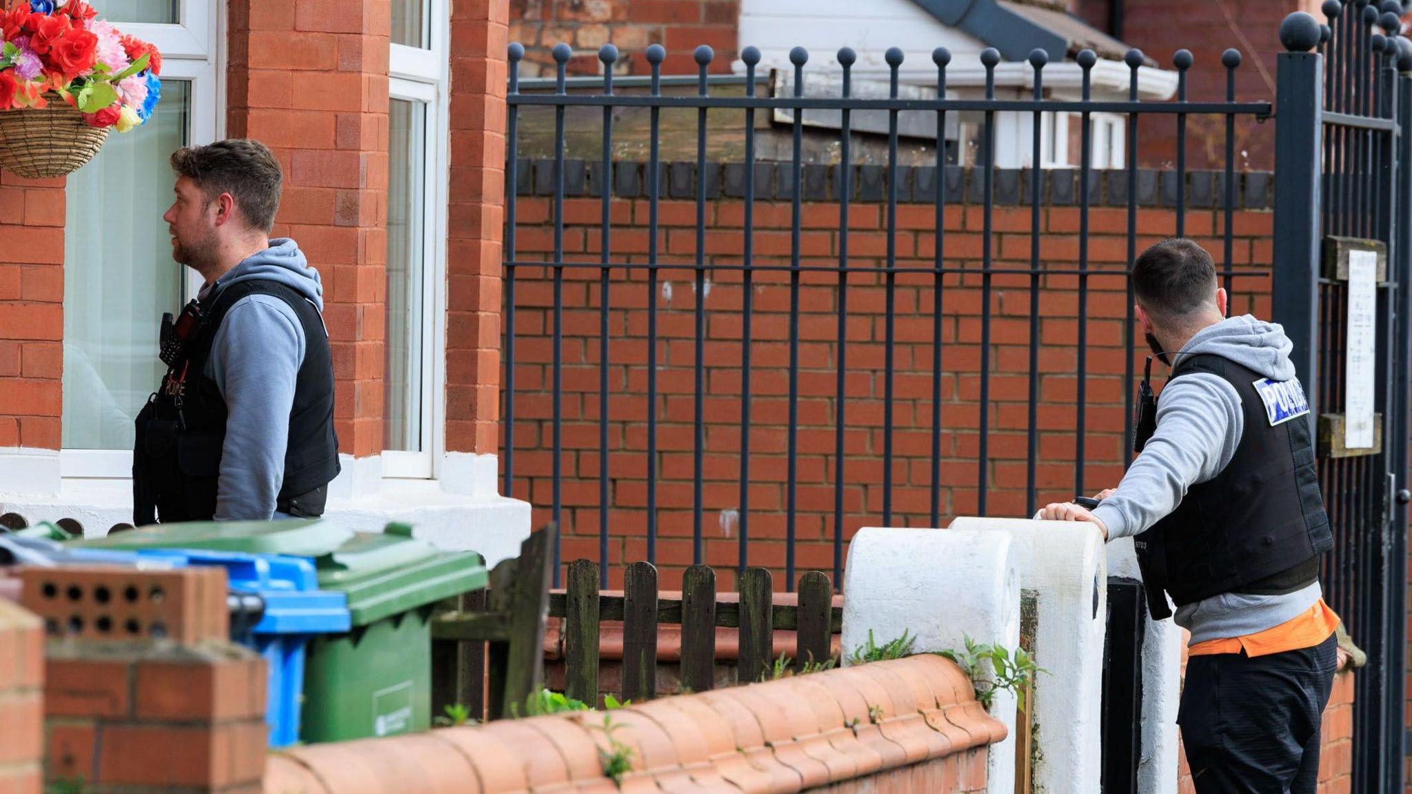 Two GMP officers in plain clothes but wearing black police vests on top stand outside a property.