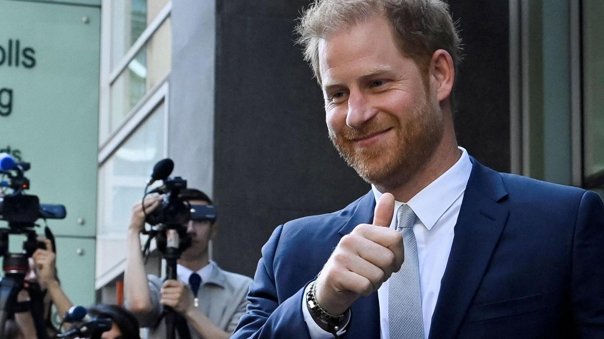 Prince Harry, dressed in a blue suit and grey tie, gives a thumbs up to supporters as he leaves the High Court's Rolls Building in 2023, with media cameras in the background, during his evidence against the Mirror Group titles who he was suing at the time for unlawful intrusion. 