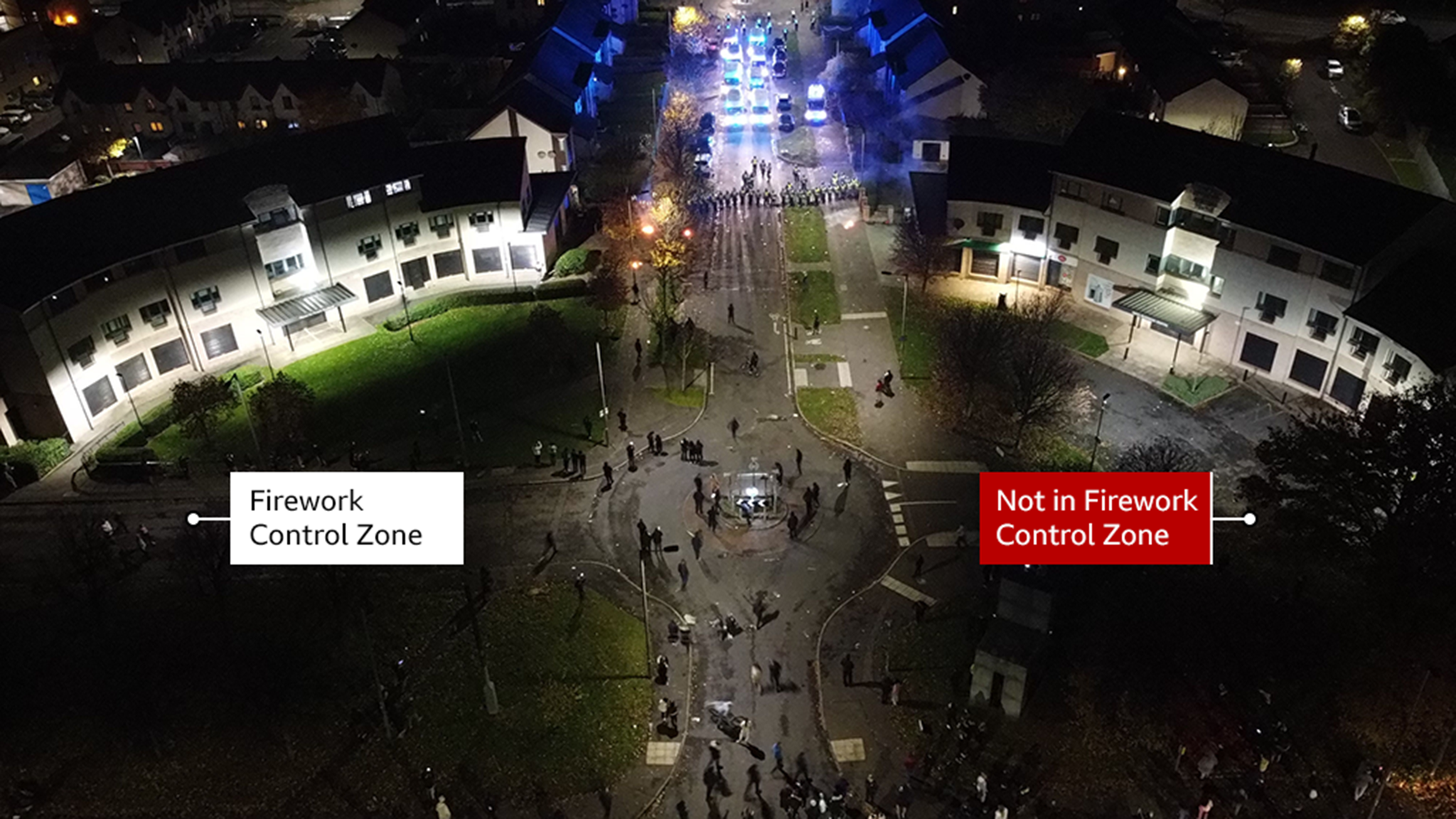 The image shows a stand off between police and young people on Hay Avenue in Niddrie on Bonfire night 2023. Police Scotland officers are standing in a line with police cars behind them. The police are blocking young people from entering the road. There are around 30 young people stood at the round about to the road.