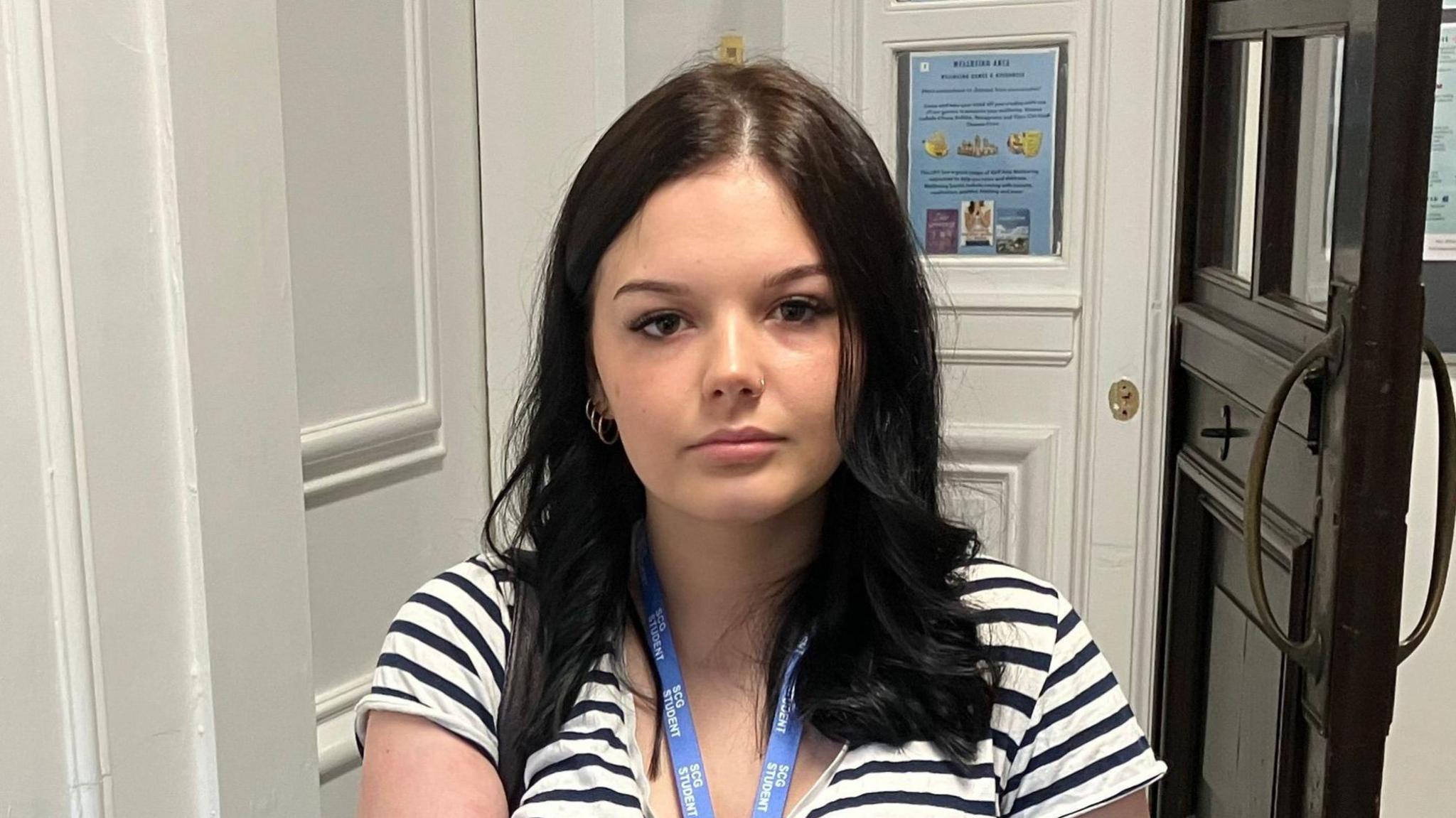 A girl wearing a white t-shirt with black stripes on it stands holding her bag and a folder. A door can be seen open behind her, with posters and leaflets on the wall of the corridor.
