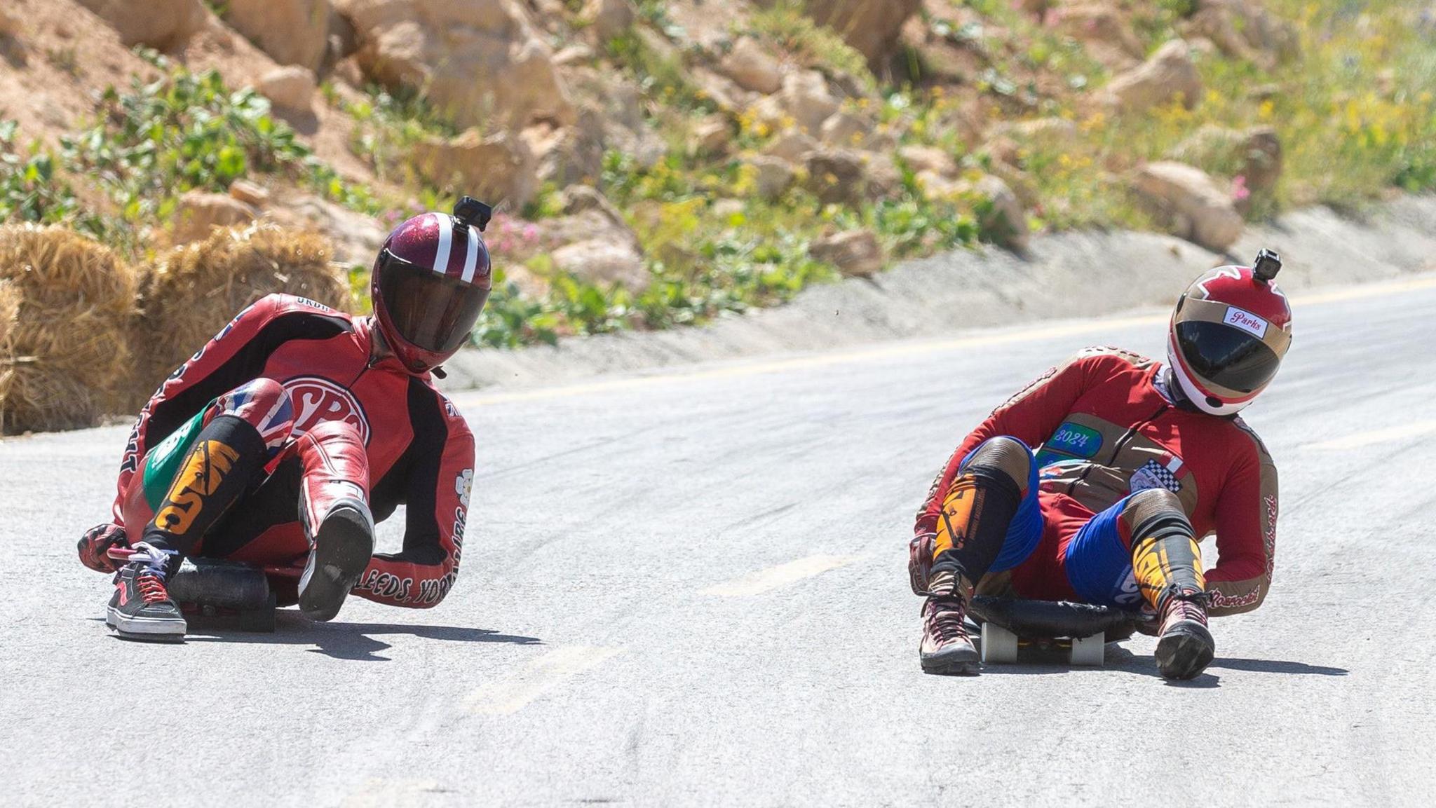Two people in brightly coloured leathers and helmets lie down on skateboards on a grey road, with a steep sandy embankment behind.