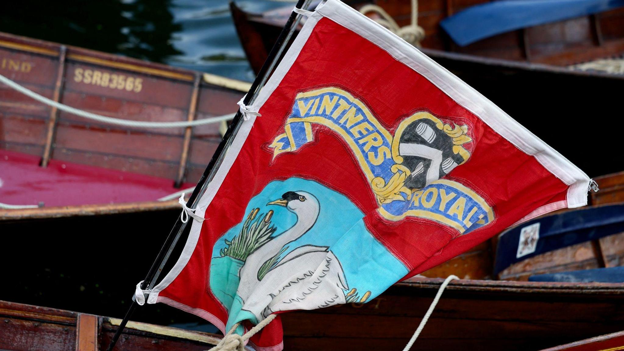 A red flag with a white border is attached to a boat docked near several wooden rowboats. The flag features a colourful design with a white swan on a blue background in the lower section, surrounded by reeds. Above the swan, a blue and gold banner reads "Vitners Royal".