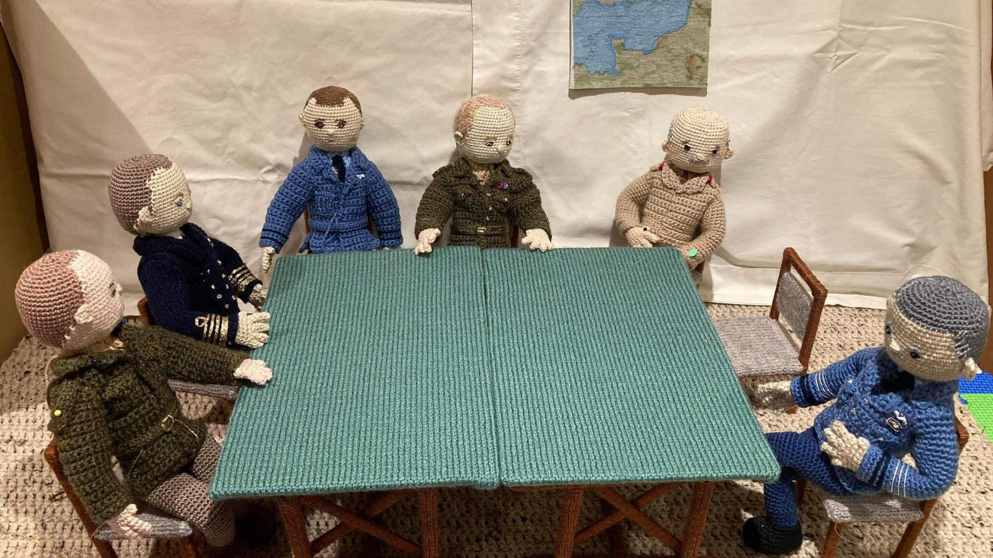 Six crochet figures are placed to look like they are sitting round a rectangular table. They each have different uniforms and outfits on. 