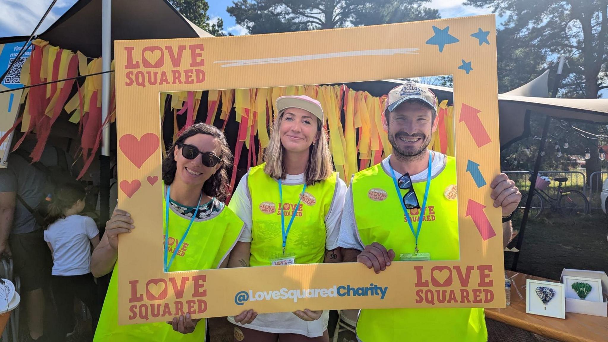 Three people from the charity Love Squared in high vis tops, holding a Love Squared sign around them.