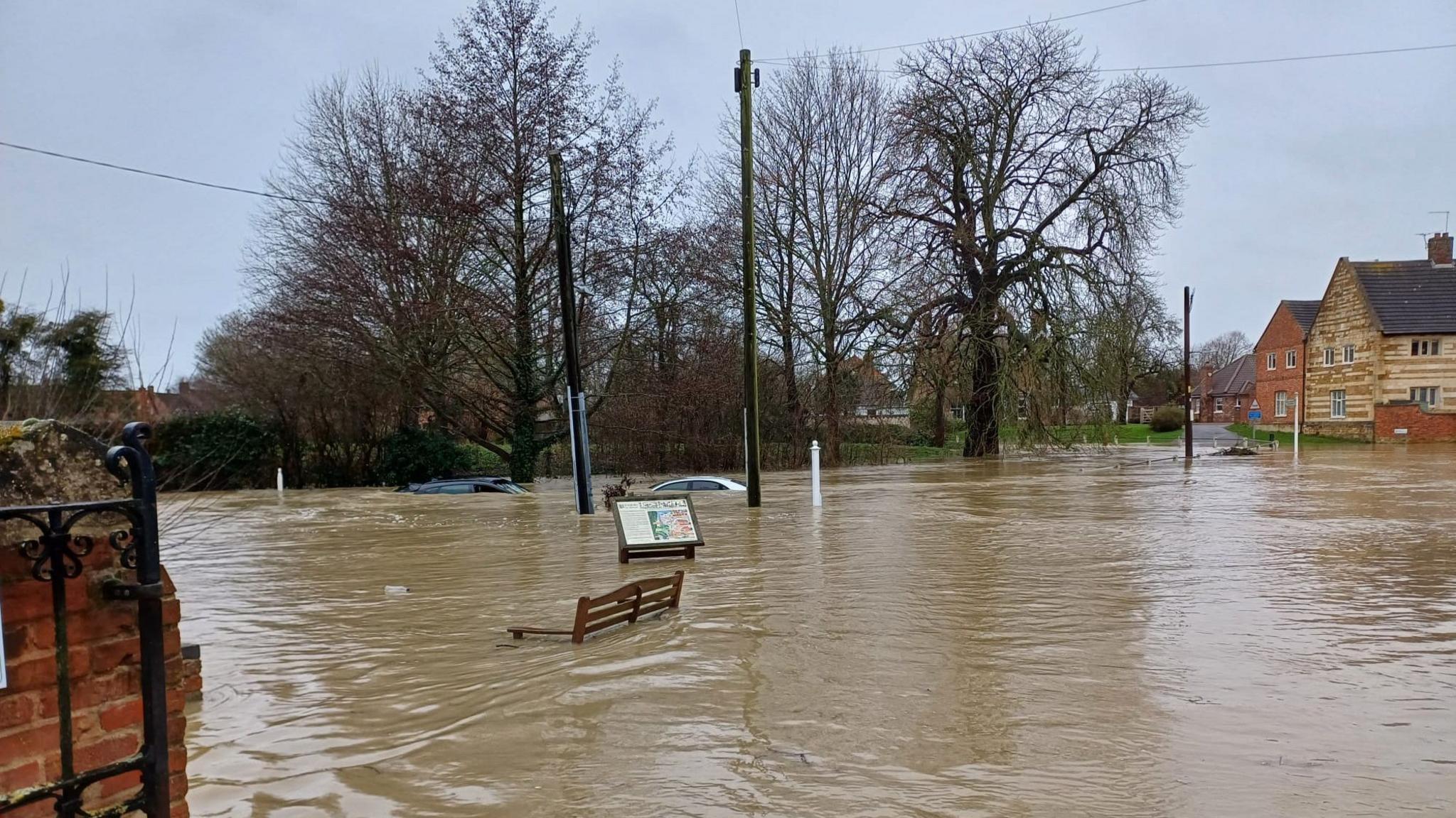 Two cars are submerged in floodwater. A bench is also partly under water.