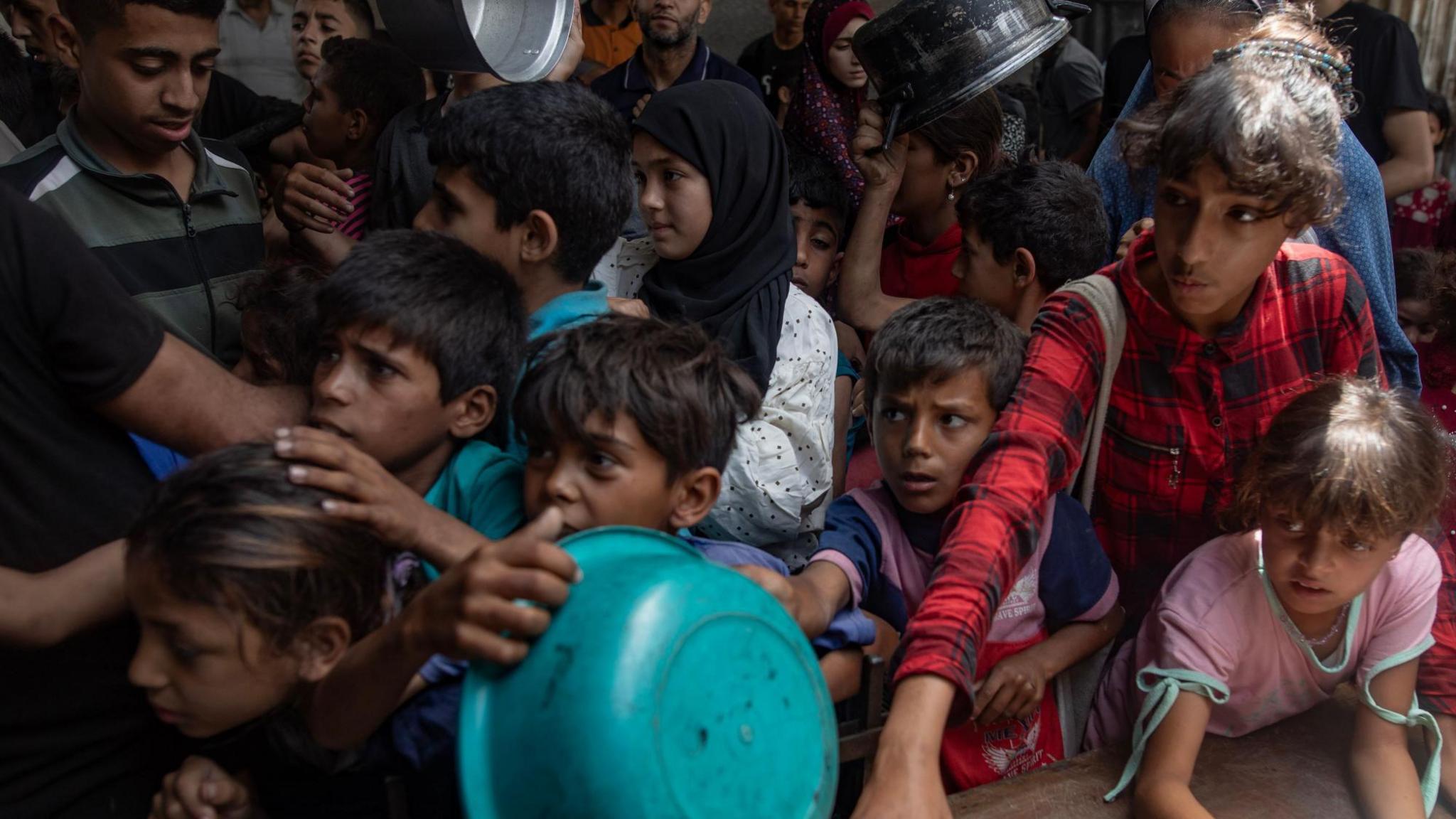 Palestinian children, some holding bowls, gather at a charity kitchen in Gaza to receive food rations.