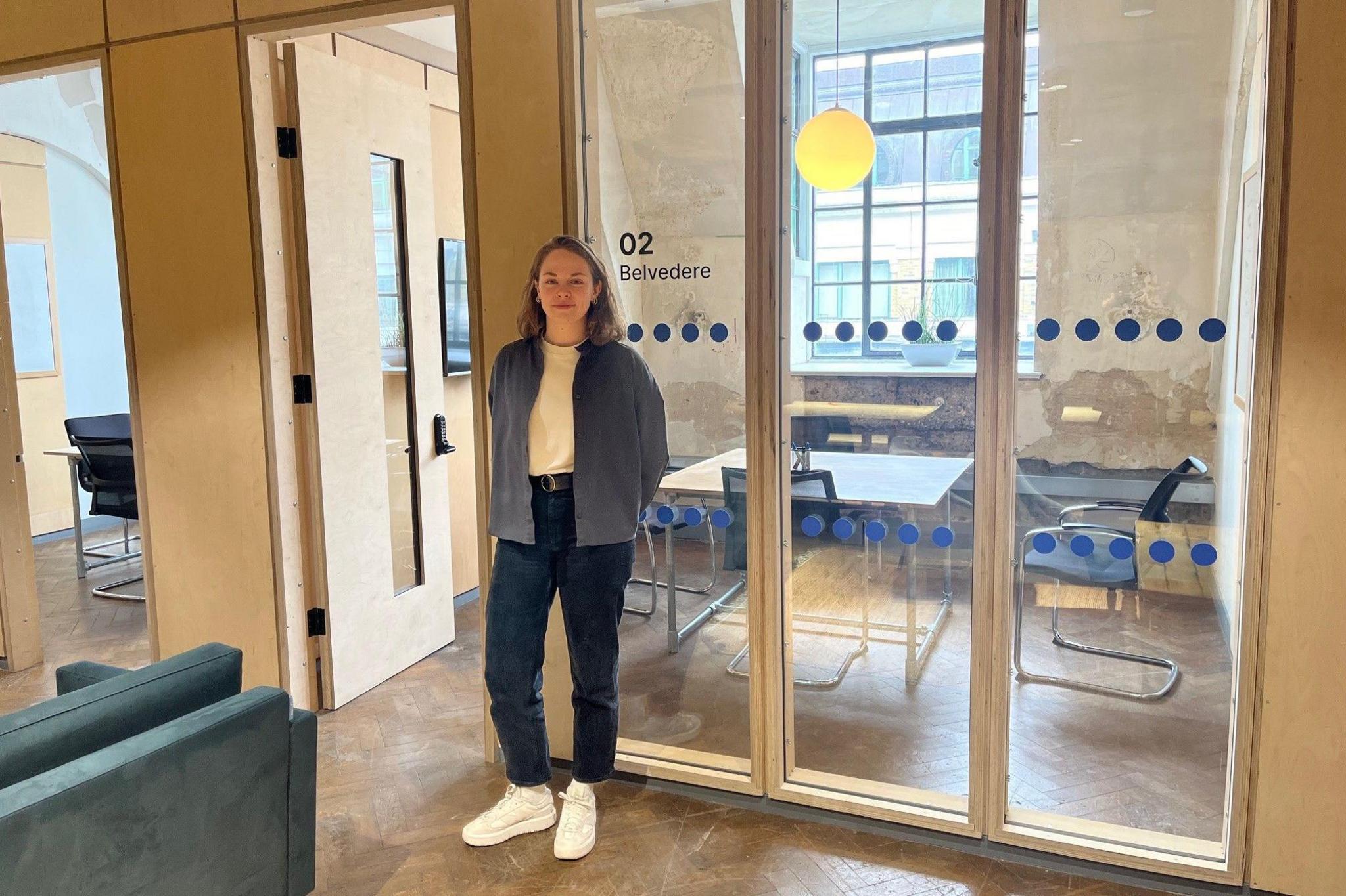 Hannah Towler wearing jeans, white T-shirt and a blue/grey top in front of an office with a table a chairs in it