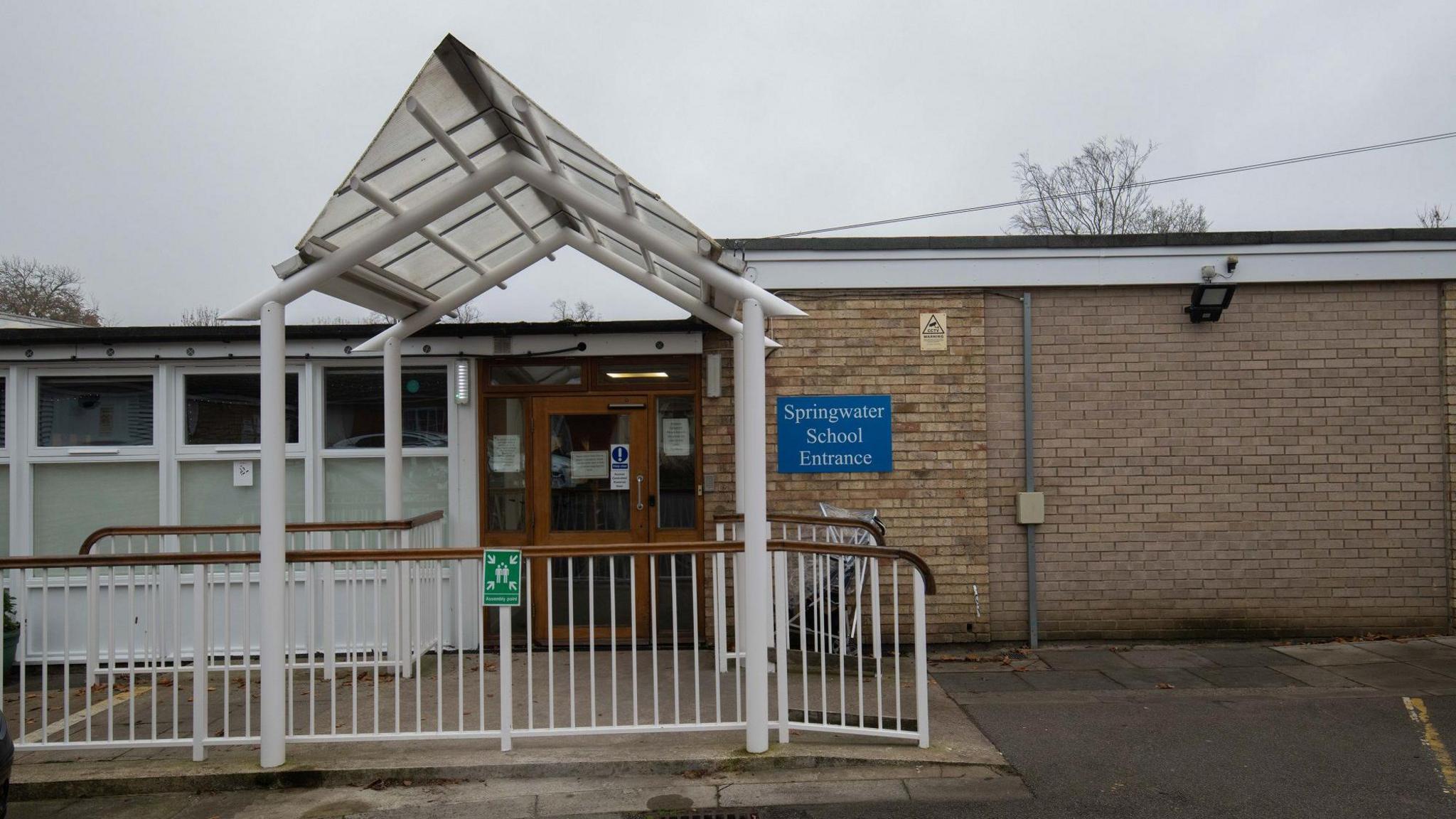 The school entrance accessed via a roofed brown automatic door. A blue sign with the words "Springwater School Entrance" is fixed to the right.