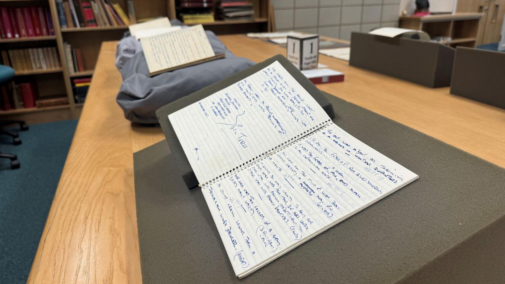 Open notebooks displayed on cushions on a wooden table. They have scribbled handwriting inside.