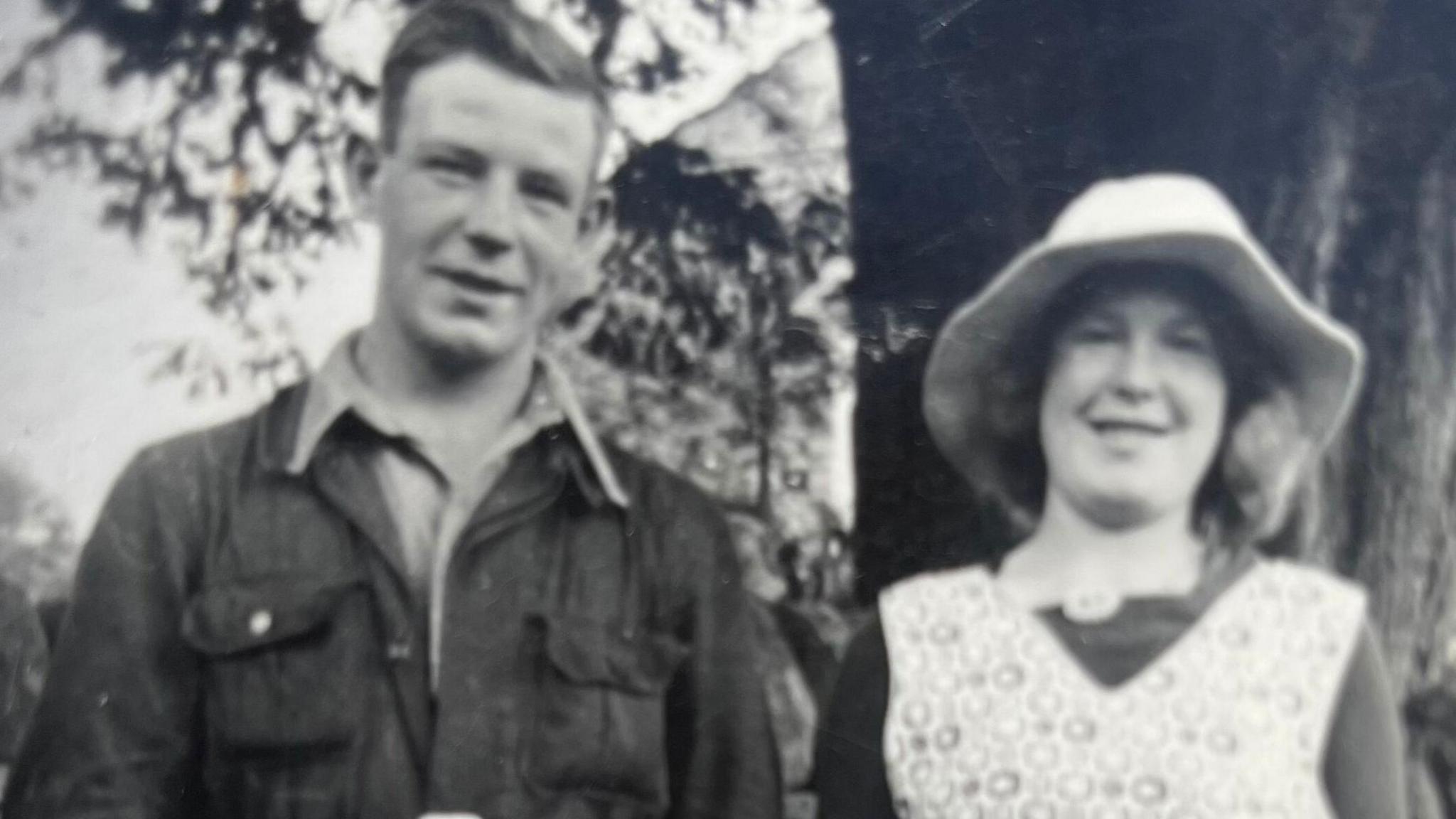 A black and white photograph of two members of Walter Renwick's family taken perhaps in the 1940s or 50s. The man on the left is wearing overalls and a shirt, the woman on the right has a pinafore and sunhat. There is a large tree visible behind them.