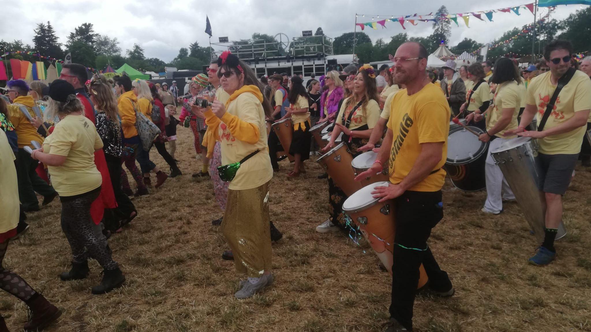 People in rows walk with samba drums through a festival site on a grey day. They are wearing yellow T-shirts, most in sunglasses.