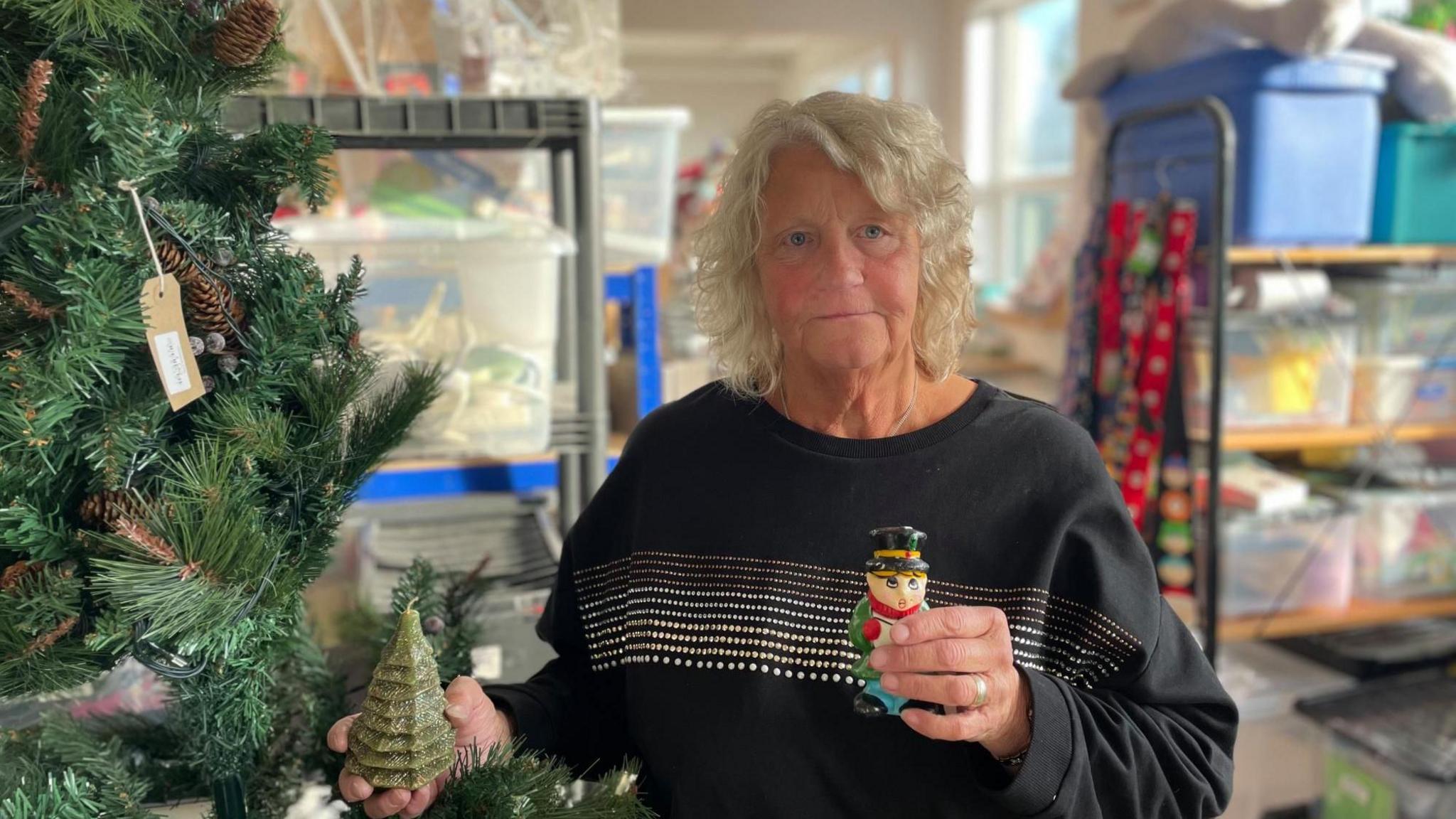 A blonde woman with a black top holds two Christmas decorations beside a Christmas tree.