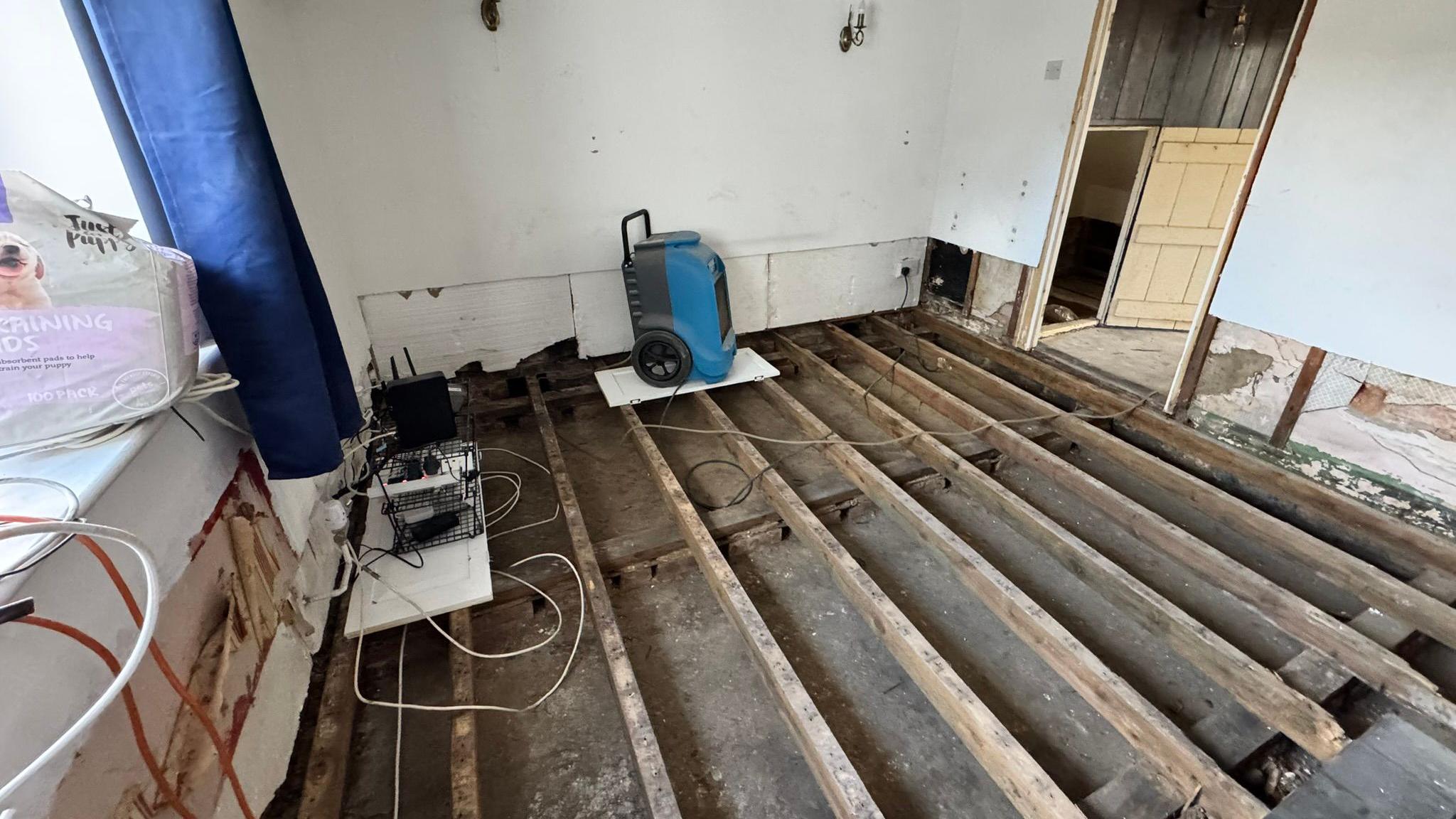 An internal room with the joists exposed in the floor and the lower part of the wall stripped back. A blue dehumidifier is plugged in. There are various wires draped around the space. The walls are white plaster and there is an open doorway on the right, with no door. A windowsill is on the left with light coming in.
