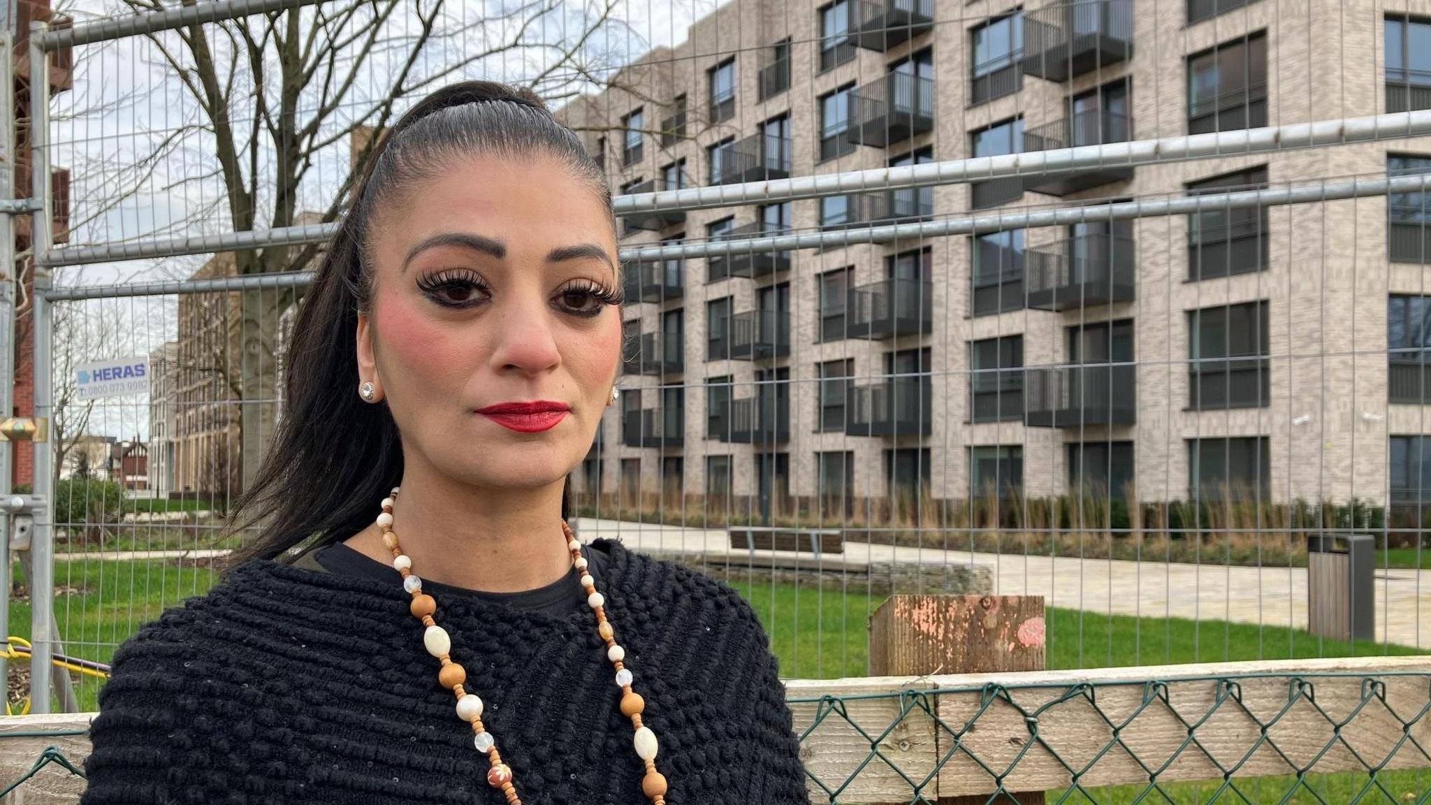 Morriam has black hair tied in a ponytail. She is wearing a black top with a white and brown wooden beaded necklace. She is standing in front of a block of new flats with a metal barrier surrounding the area.