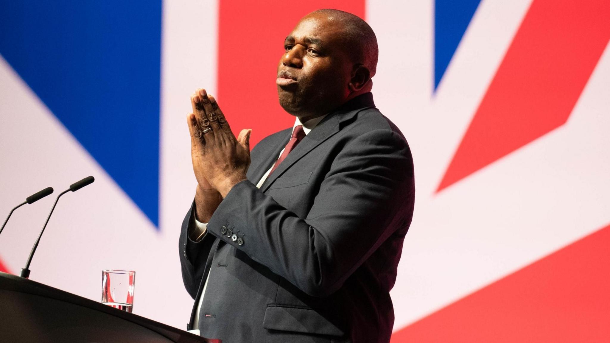 Foreign Secretary David Lammy speaking during the Labour Party Conference in Liverpool. He is at a lectern with microphones and is wearing a grey suit, white shirt and red tie. Behind him is a union flag.