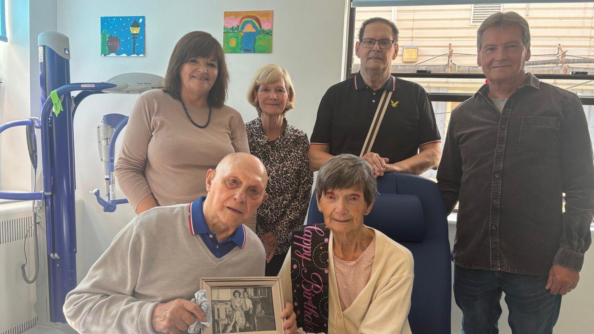 Elizabeth Ross, an elderly woman sits on hospital chair. Her husband John Ross, an elderly man, crouches beside her. Their four children stand behind them.  