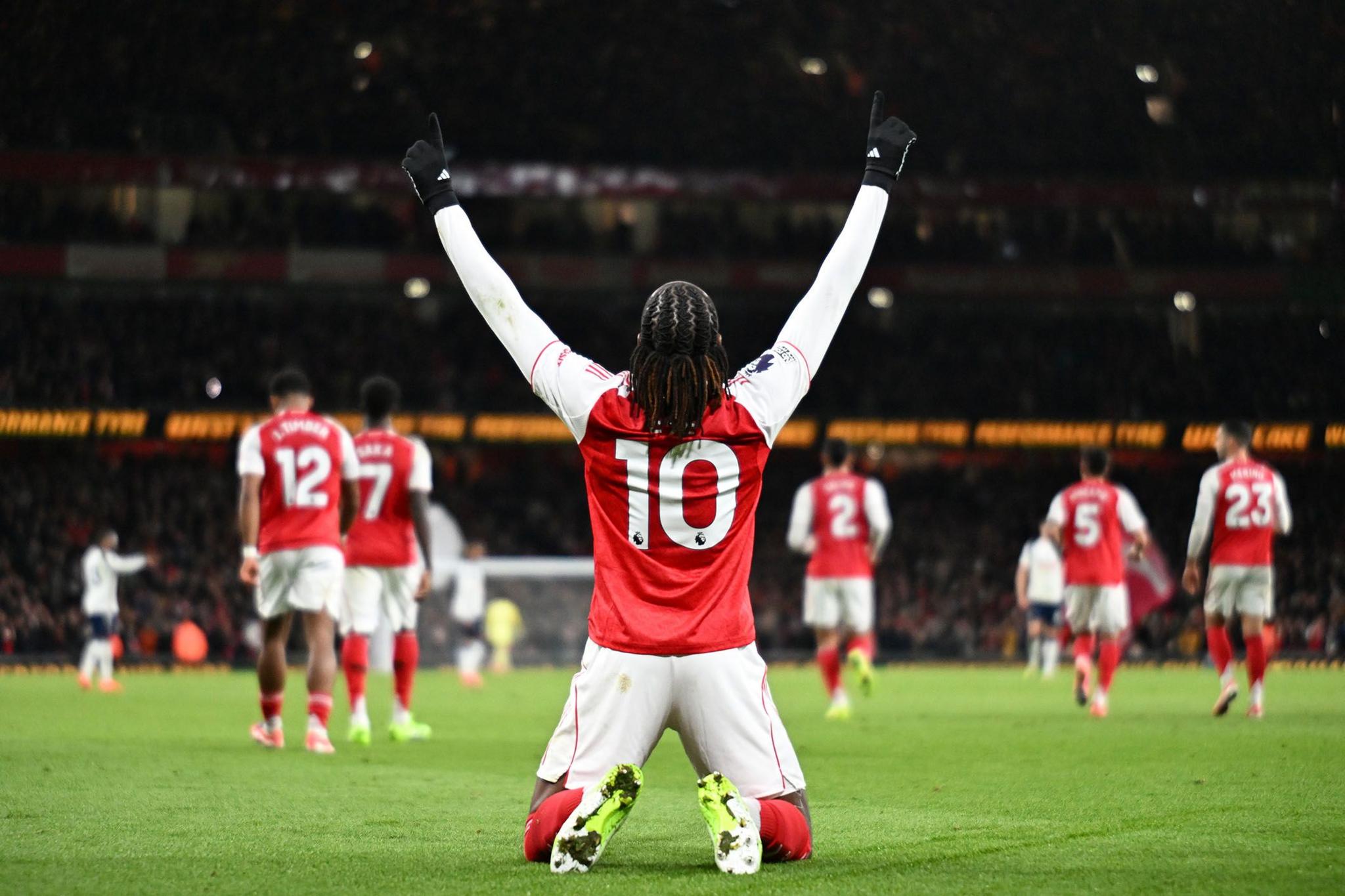 Football player wearing a red and white jersey with number 10 kneeling on the pitch, arms raised in celebration, with team-mates and opponents visible in the background under stadium lights.