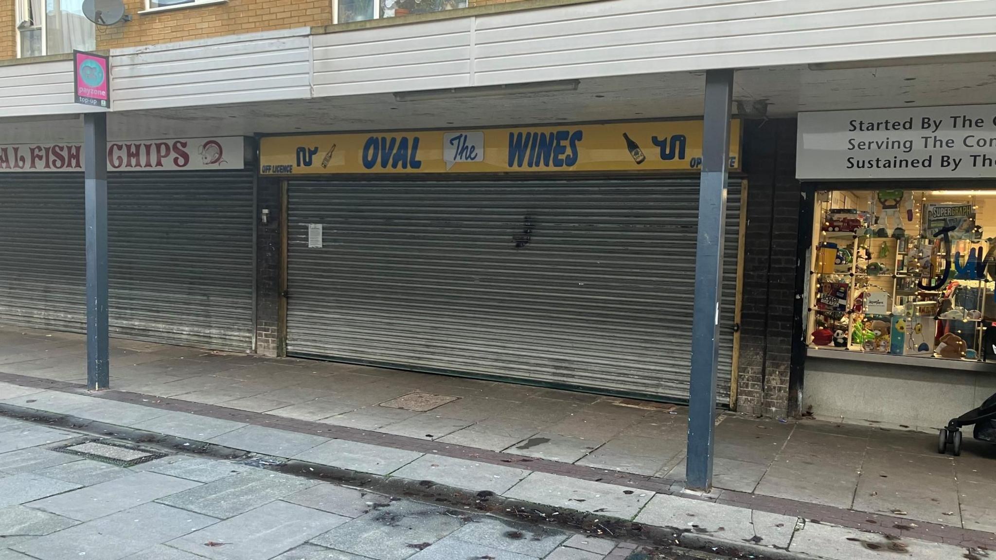 Exterior shot of Oval Wines in Stevenage. It has a blue and yellow sign above metal shutters covering the whole of the front of the store, which is on a row of shops in a shopping precinct.