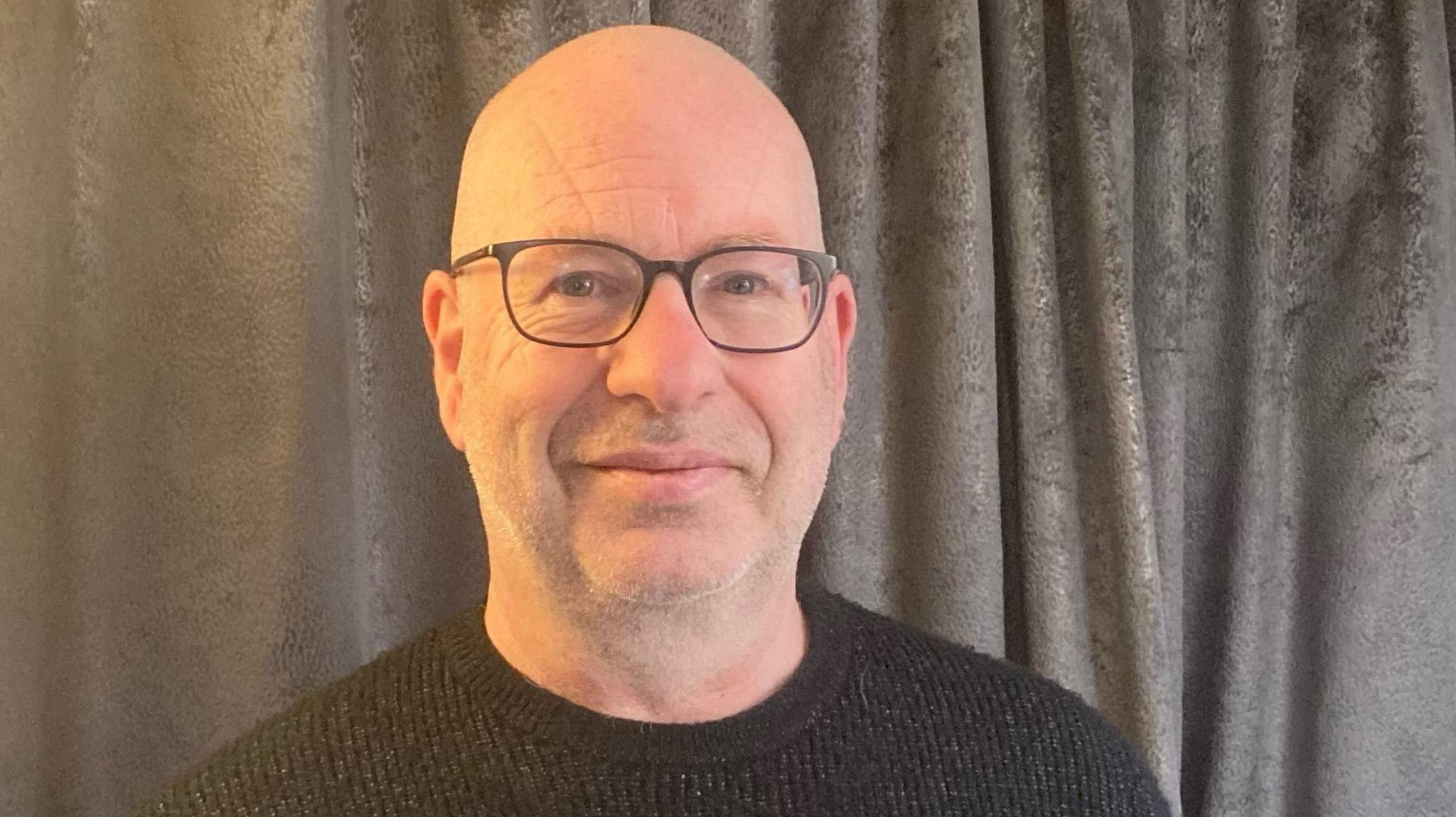 Davy Uprichard wearing a grey jumper and glasses looking into the camera with a slight smile. He is standing in front of a grey curtain