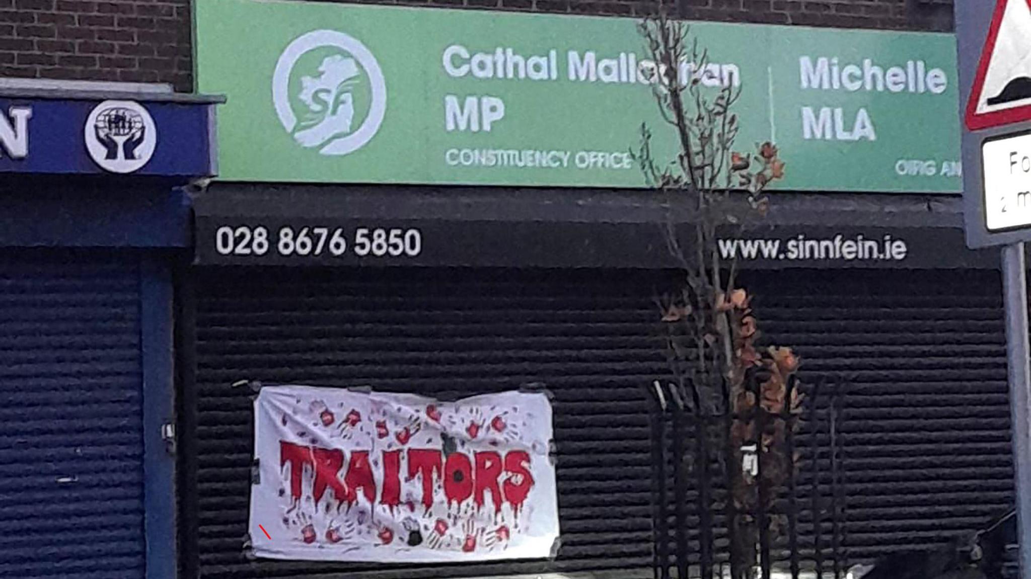 A wide shot of the office of Michelle O'Neill and Cathal Mallaghan. A green sign bears their names and the Sinn Féin party logo. Below it are black shutters, which a banner is stuck to. It reads "Traitors" in red writing made to look like blood and with bloody hand prints. The "O" is styled as a Remembrance poppy.