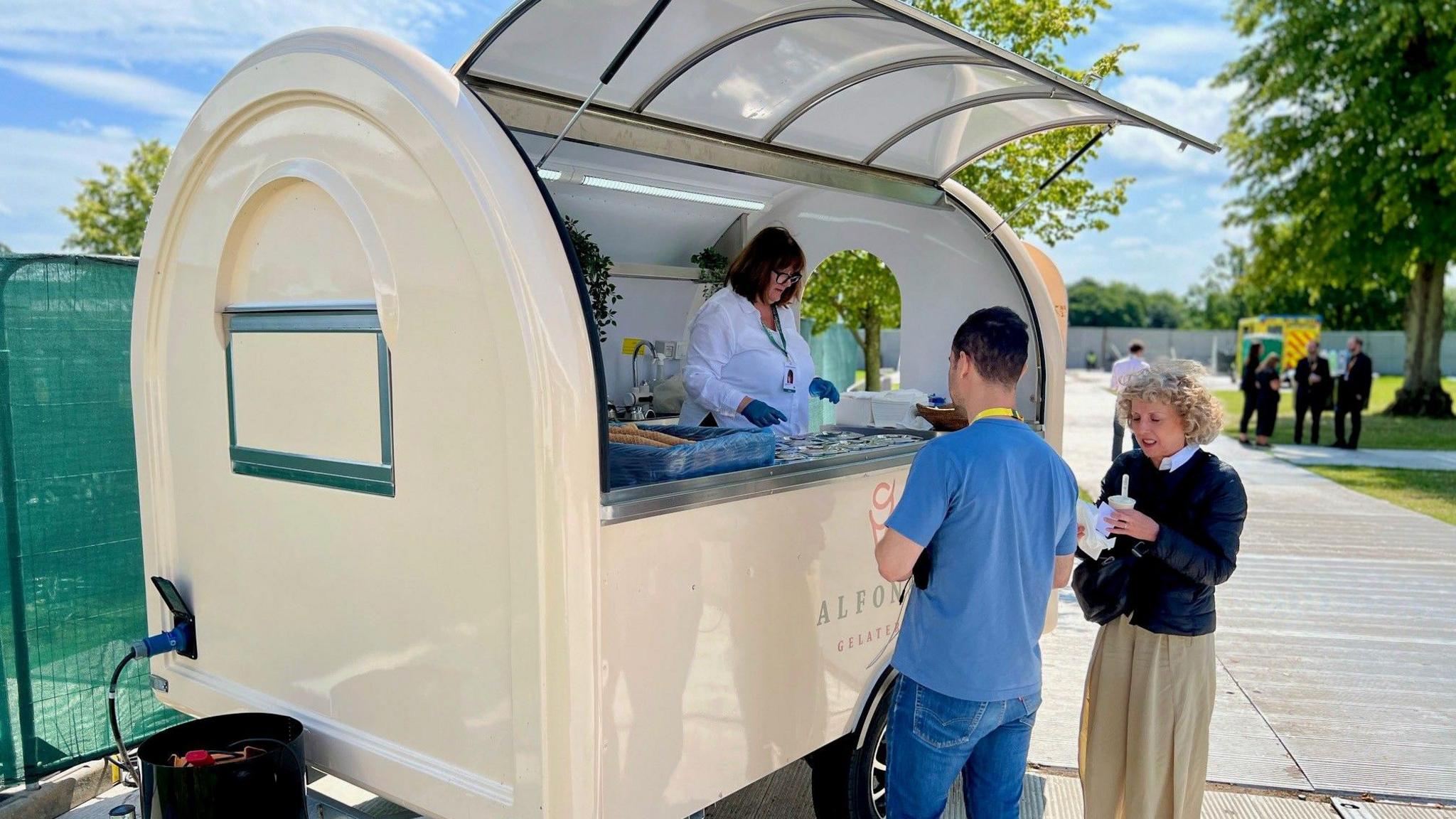 A man and woman stand next to a ice cream kiosk where it appears they are being served by a woman behind the counter