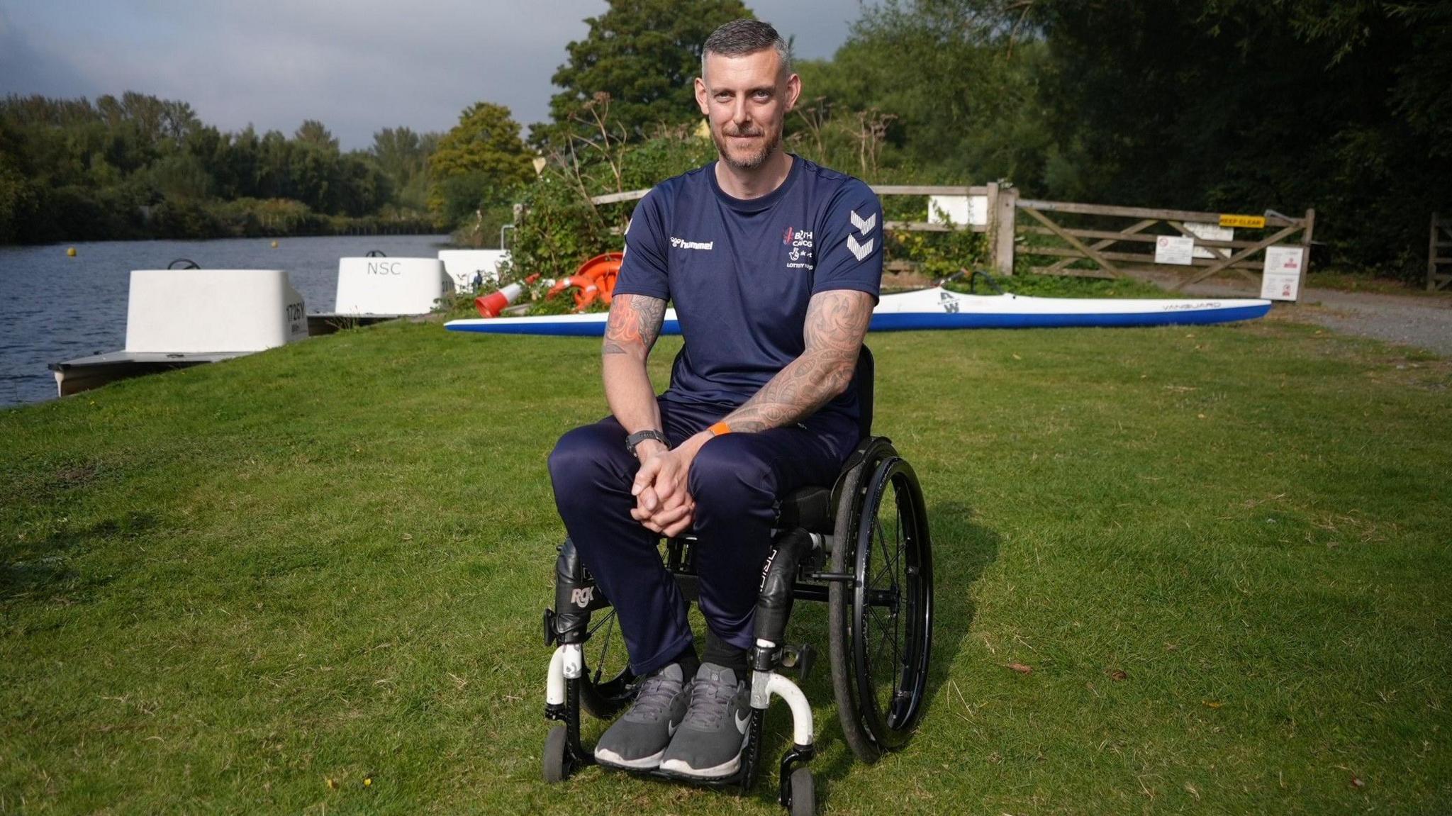 A smiling Shaun Cook looks directly at the camera. He uses a wheelchair and is photographed in his blue British Canoeing tracksuit. He is positioned on a riverbank by a body of water.