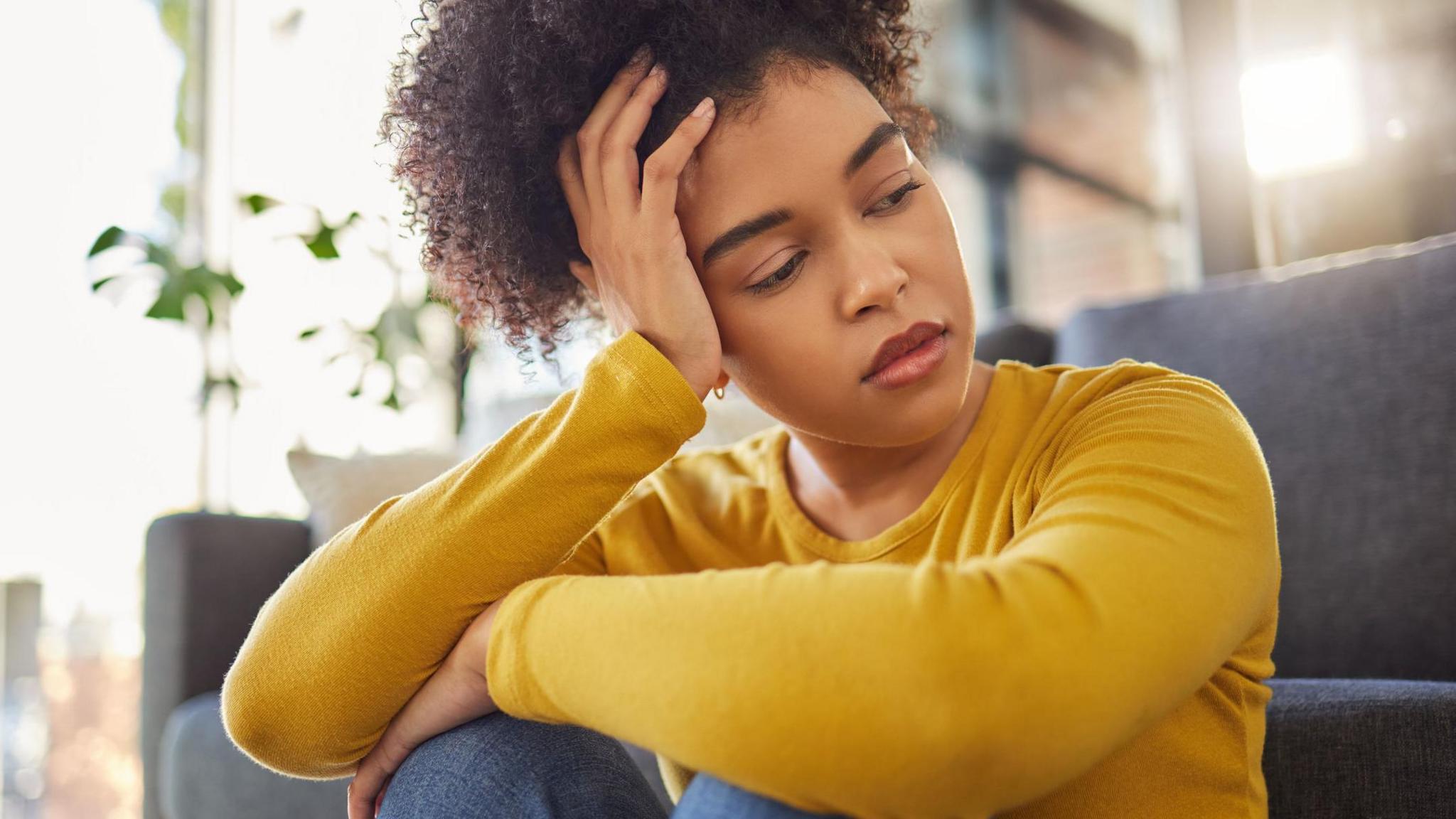 Stock image of a woman looking sad and thoughtful, sitting on the floor by a blue sofa with her knees hitched up and her left arm across them. She is holding her head with her right hand. She is wearing a yellow top and blue jeans. 