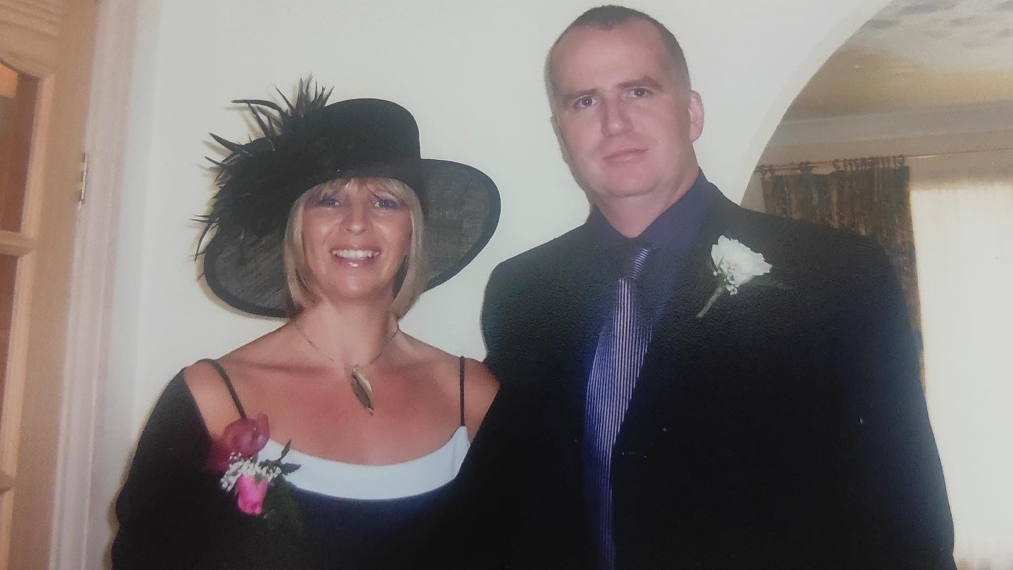 Chris and Jane when they first started fostering. Jane is wearing a black top and hat, is smiling at the camera and standing next to Chris, who is wearing a black shirt and blue shirt and tie. They both have flowers pinned to them, as though they are attending a wedding.