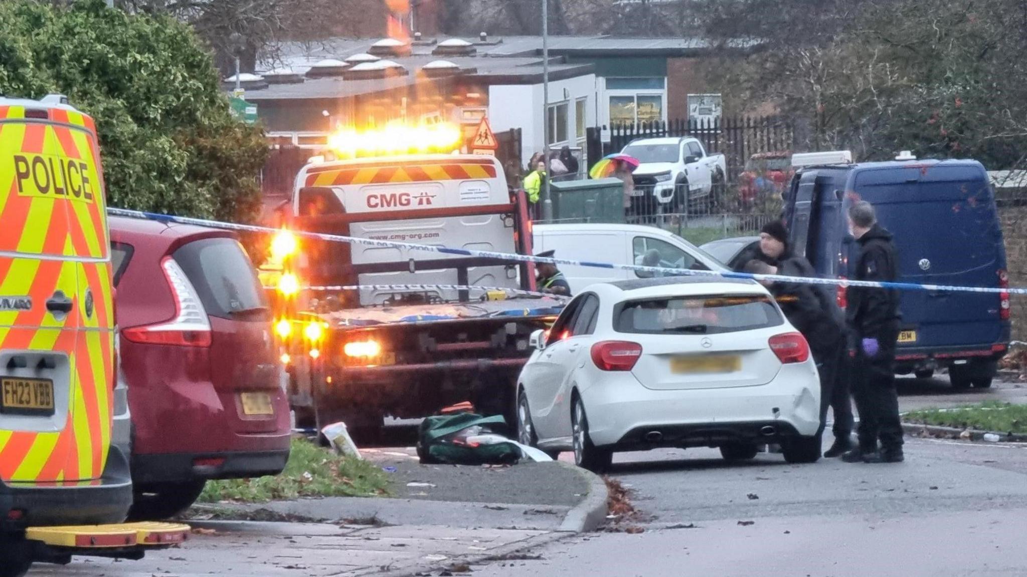 A police van, a maroon people carrier, a car removal truck and a white car parked in a estate. Several police officers are talking to each other and police tape has been set up.