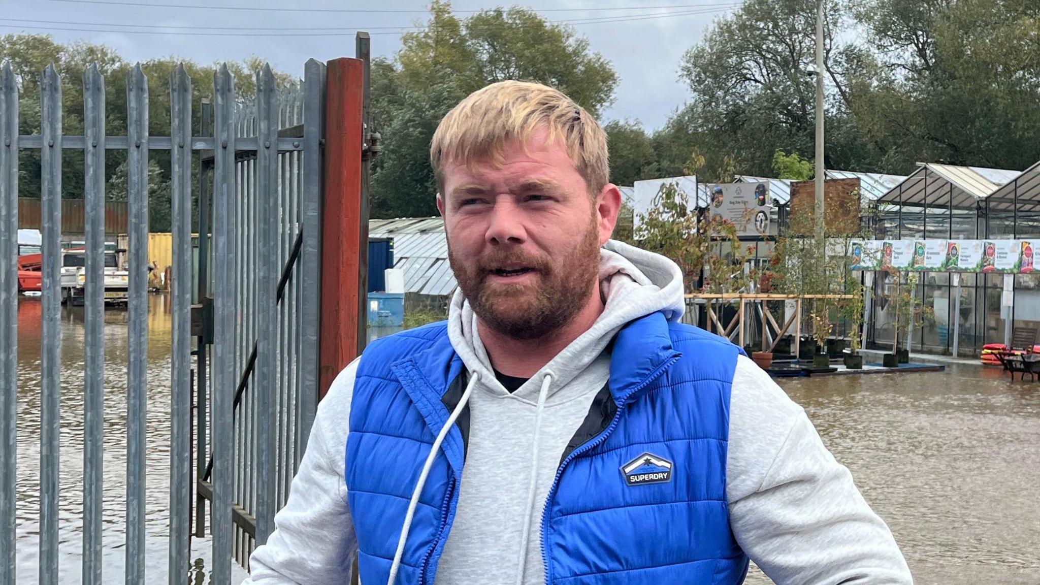 Kyle Nester, looking off camera, standing in front of flood water. He is wearing a blue gilet over a light grey hooded top. He has a light brown beard and blond hair. Behind him is a large body of water, surrounding glass houses and young trees inside a garden centre setting. A heavy metal fence and gate are visible on the left of the picture. There are trees and dark skies in the far background. 