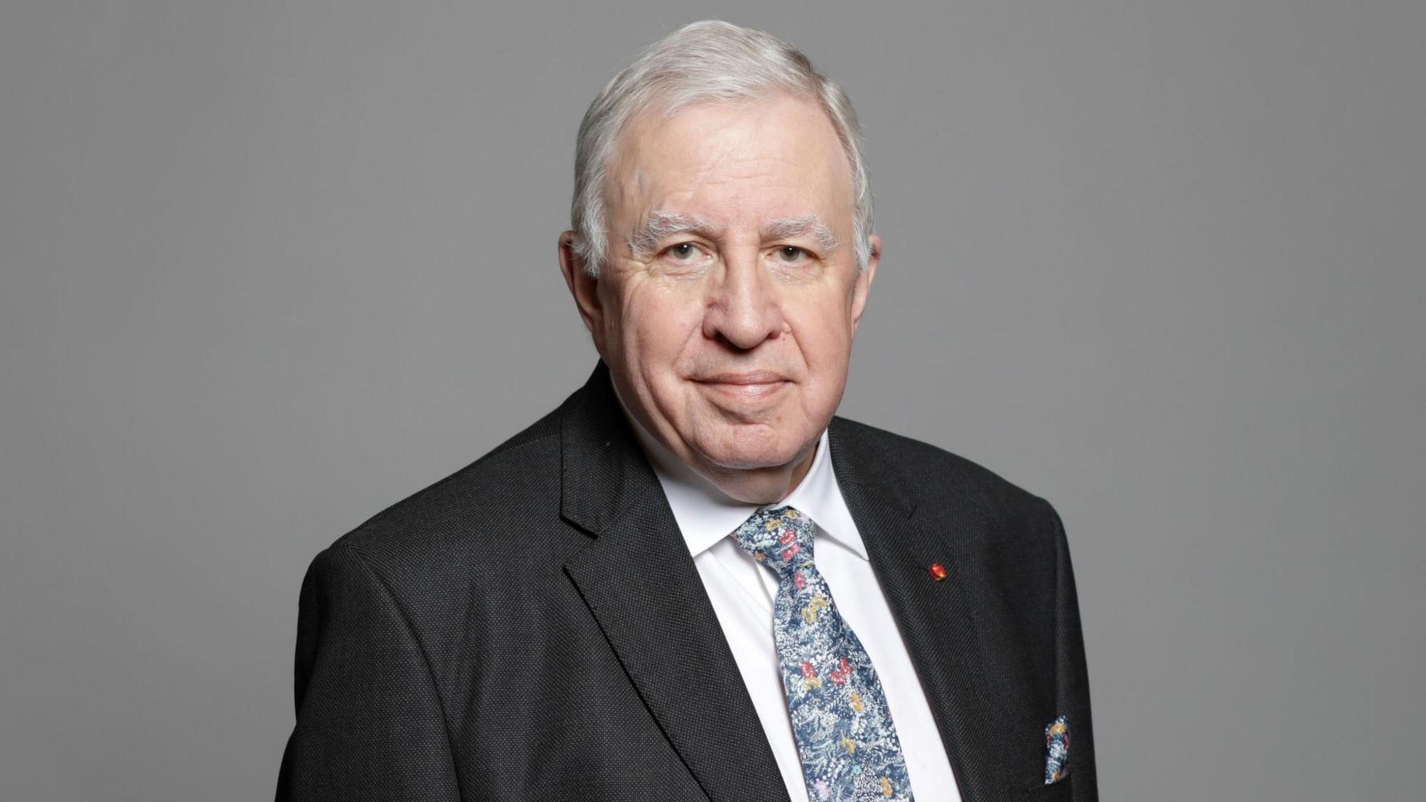 An official portrait of Lord Paul Murphy - a man with grey hair wearing a black suit jacket, a white collared shirt and a blue patterned tie smiles at the camera while standing against a grey background.