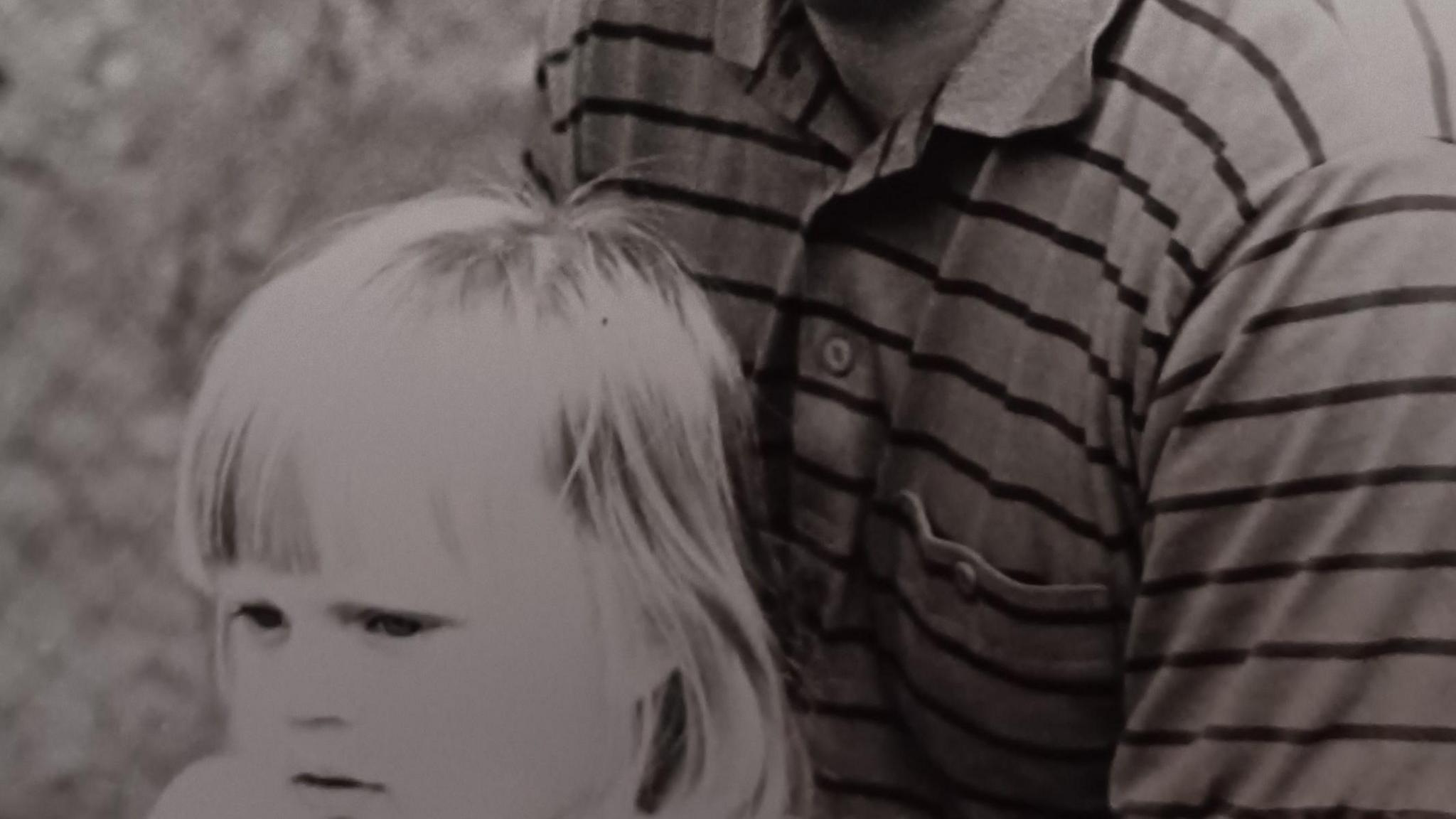 A black and white image of Alice as a child, looking off into the distance. She is sitting on Andrew's lap, who is cut out of the image so only his chest is seen. 