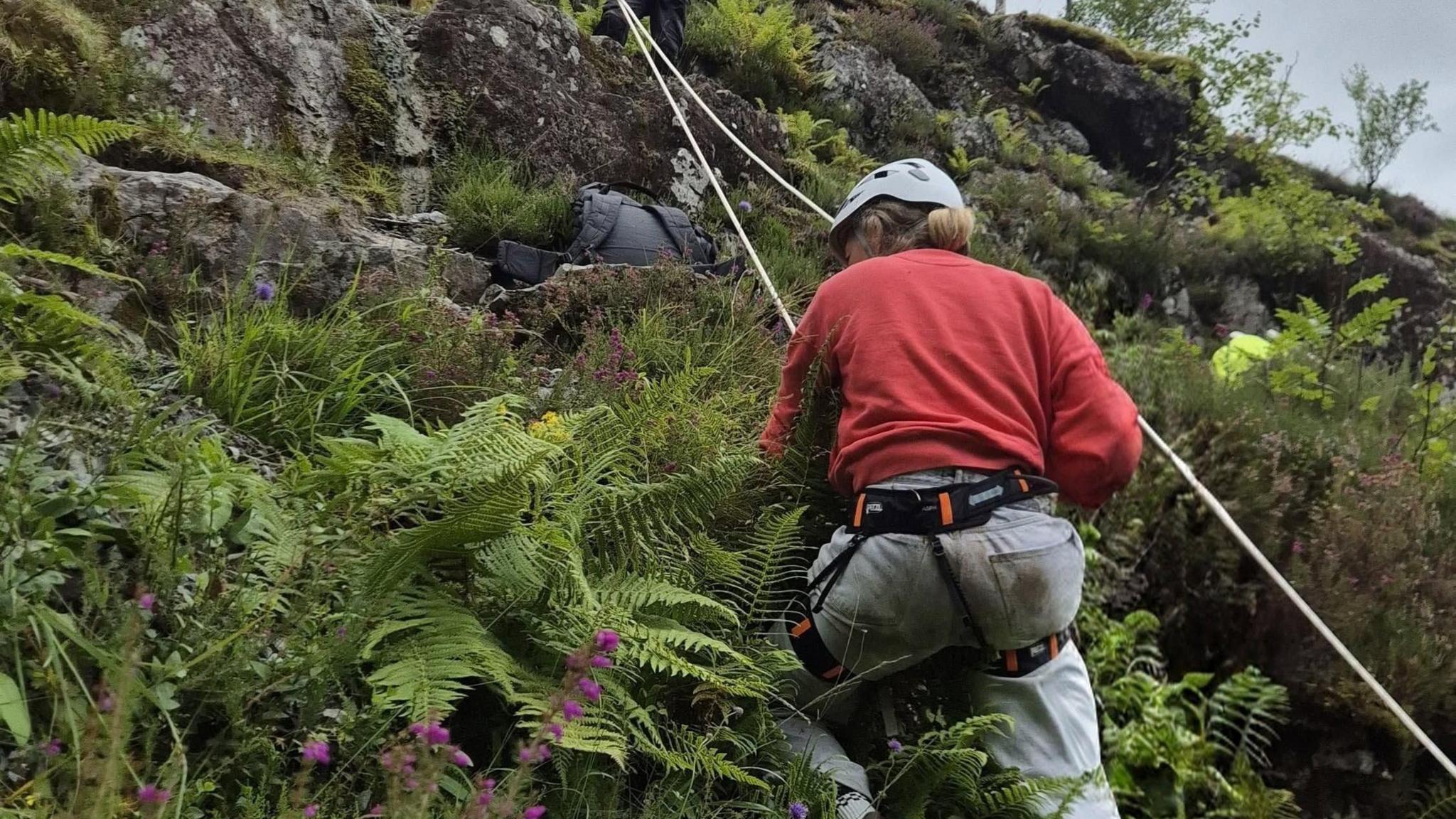 A woman in red jumper and grey trousers on ropes down the side of a hill in Glencoe.