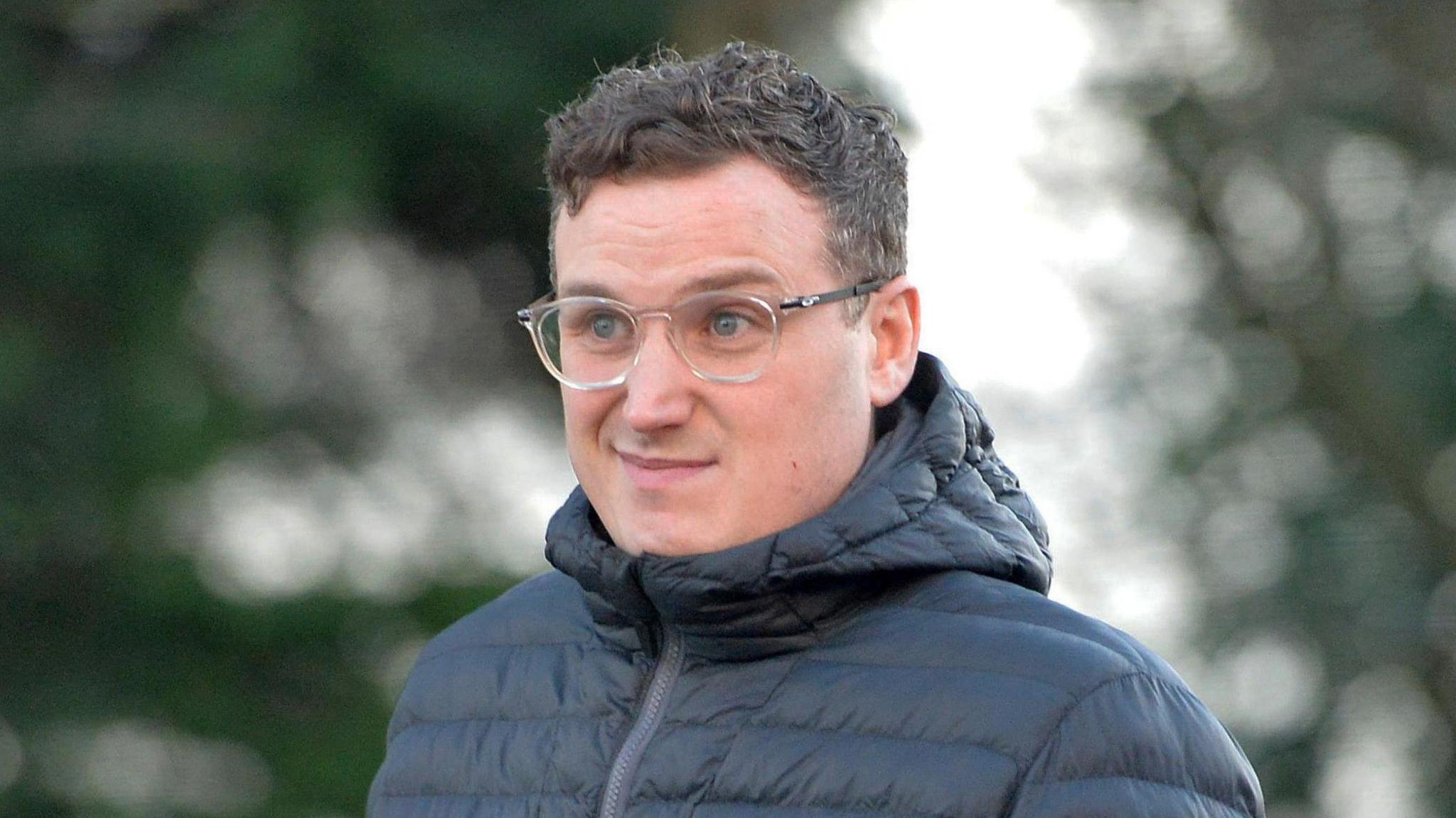 A man with dark hair and clear framed glasses. He has blue eyes and is wearing a black puffer jacket.