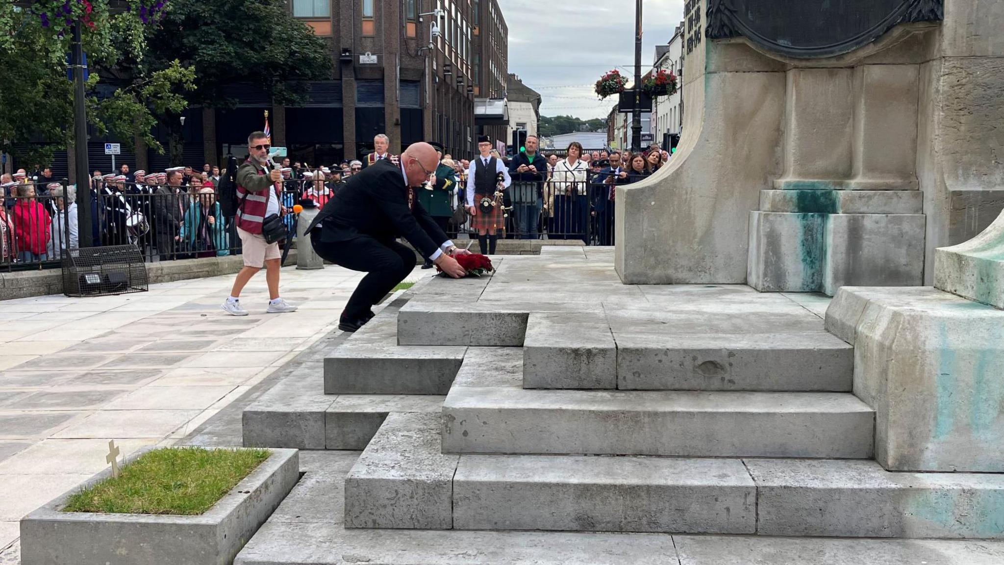 An older man wearing a black suit is bent over in front of a large stone war memorial. He is placing a poppy wreath on one the memorial's steps. There's a large crowd in the background watching him.