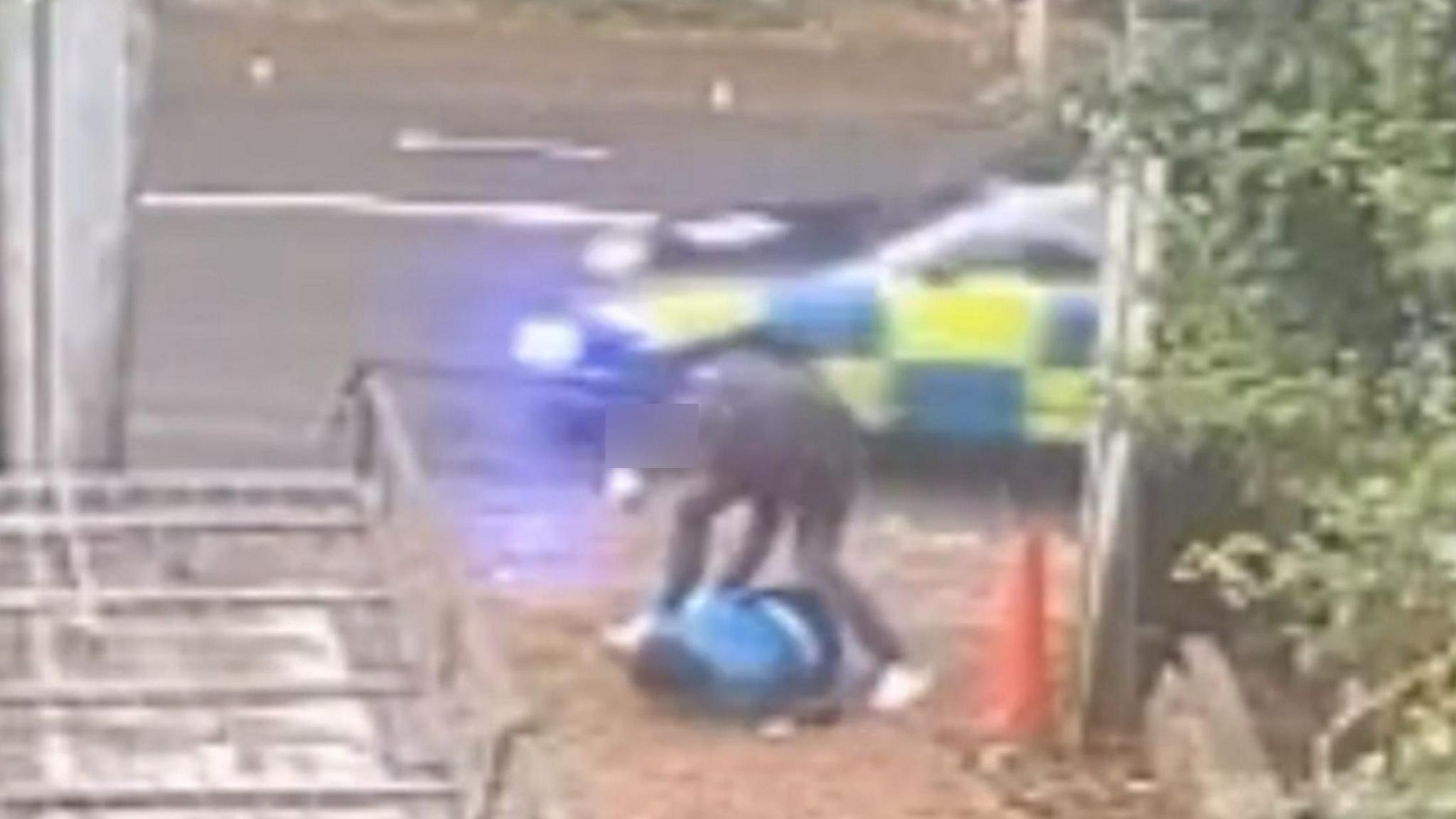 CCTV footage showing a man in dark clothing with his right fist raised crouching over a man in a blue jacket who is face down on the ground.  There is a wooden gate to the right and a police car is passing on the road behind