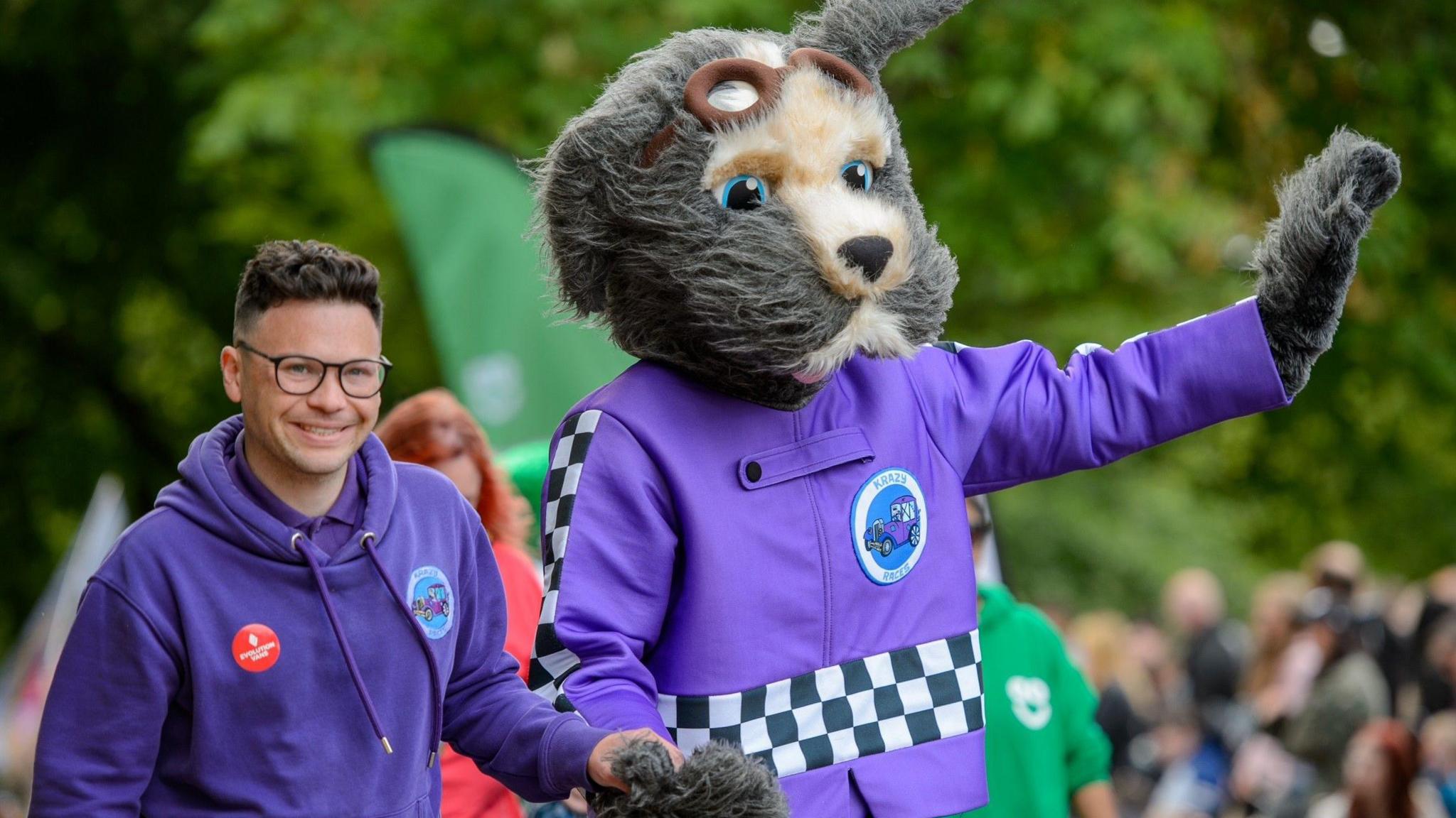 A man walking down a street in a purple hoodie. He is holding the hand of a fancy dress character. The character is a grey and white dog that is dressed in purple mechanics overalls. The character is waving to the crowd.