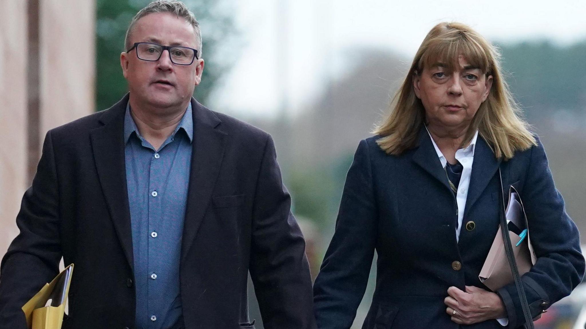 Stuart and Linda Allan walking down a road hand in hand towards the camera. He is wearing blue jeans, a blue shirt and a dark jacket and she she is wearing blue jeans and a blue jacket over a white shirt. She has a handbag over her shoulder and they are both carrying folders.